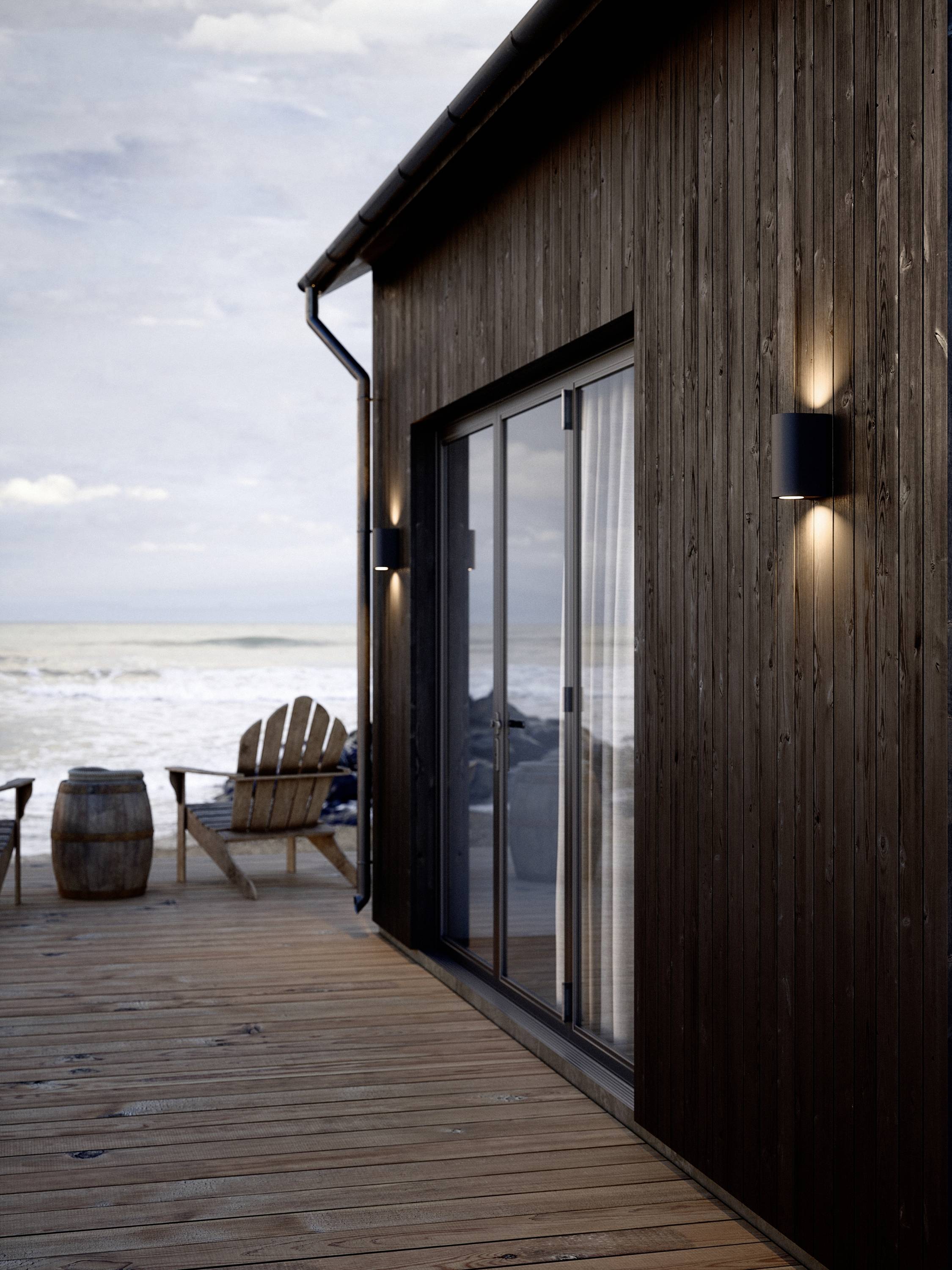 Ein Holzdeck mit Meerblick. Zwei Holzstühle und ein Fass stehen neben einer modernen Holzwand mit Glas-Schiebetüren.