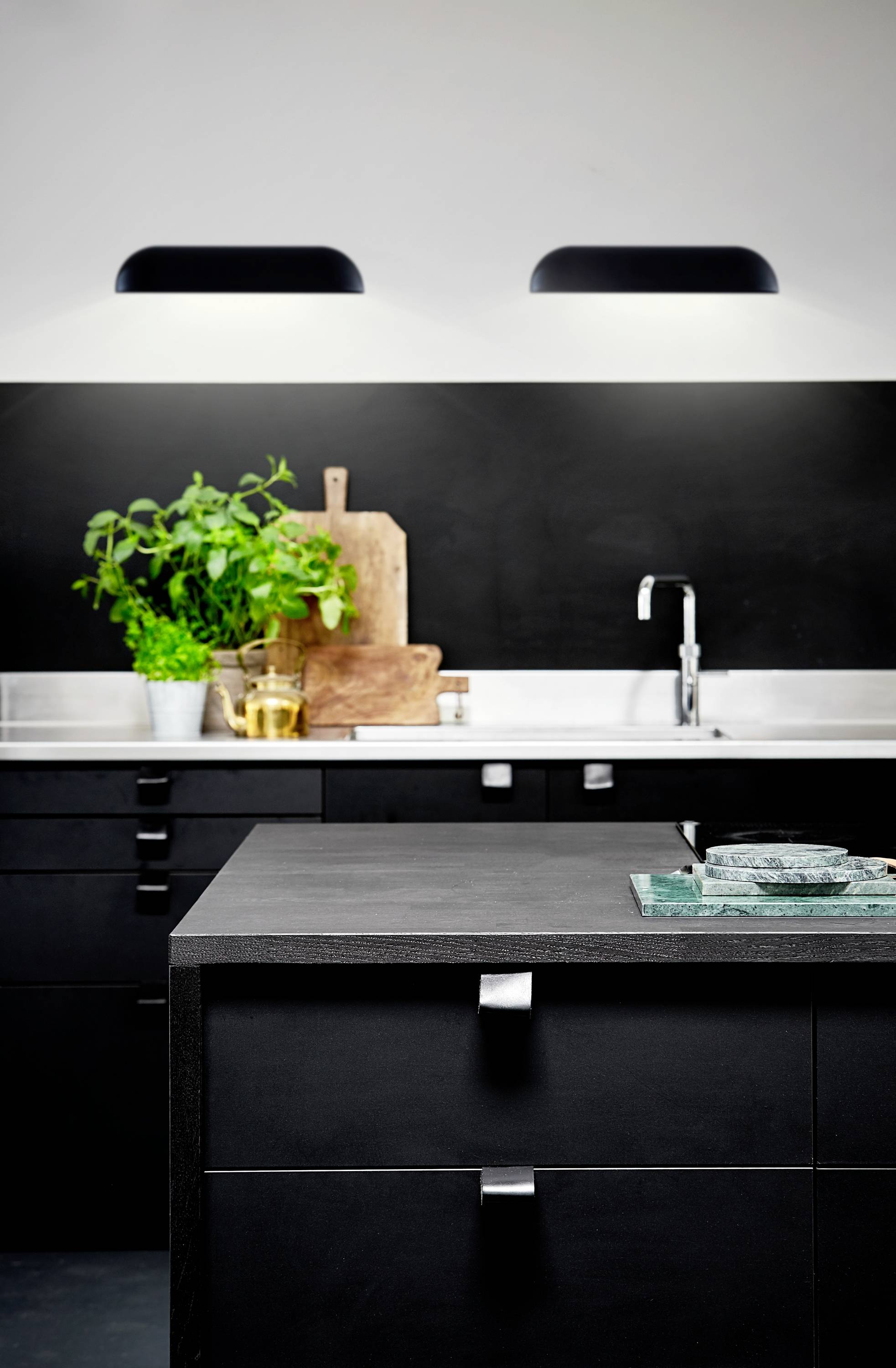 Moderne Küche mit schwarzen Schränken, Edelstahlspüle, Schneidebrettern und frischen Kräutern auf der Arbeitsplatte.
