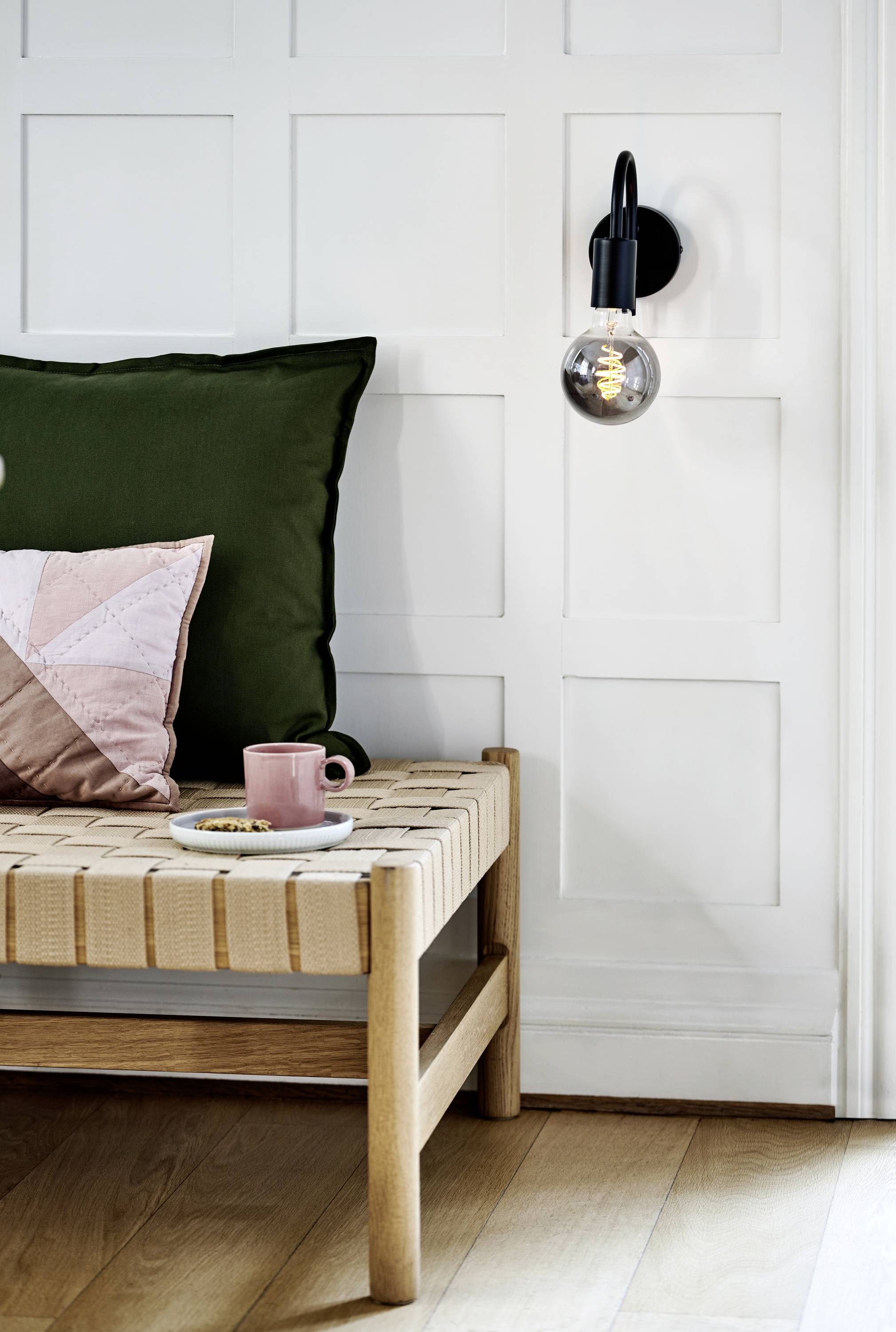 Holzbank mit grünen und rosa Kissen steht vor einer weißen Wand mit Paneelen. Eine schwarze Wandlampe ist eingeschaltet. Ein Tisch enthält eine rosa Tasse.