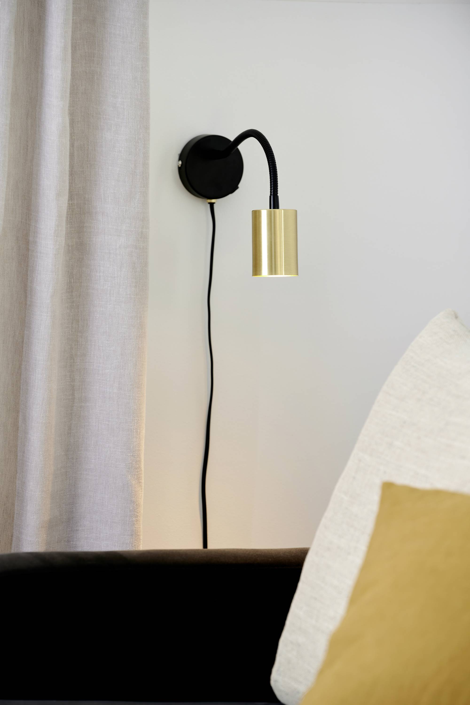 Wandleuchte mit goldfarbenem Lampenschirm, montiert an einer weißen Wand neben beigen Vorhängen und einem Sofa mit Kissen.