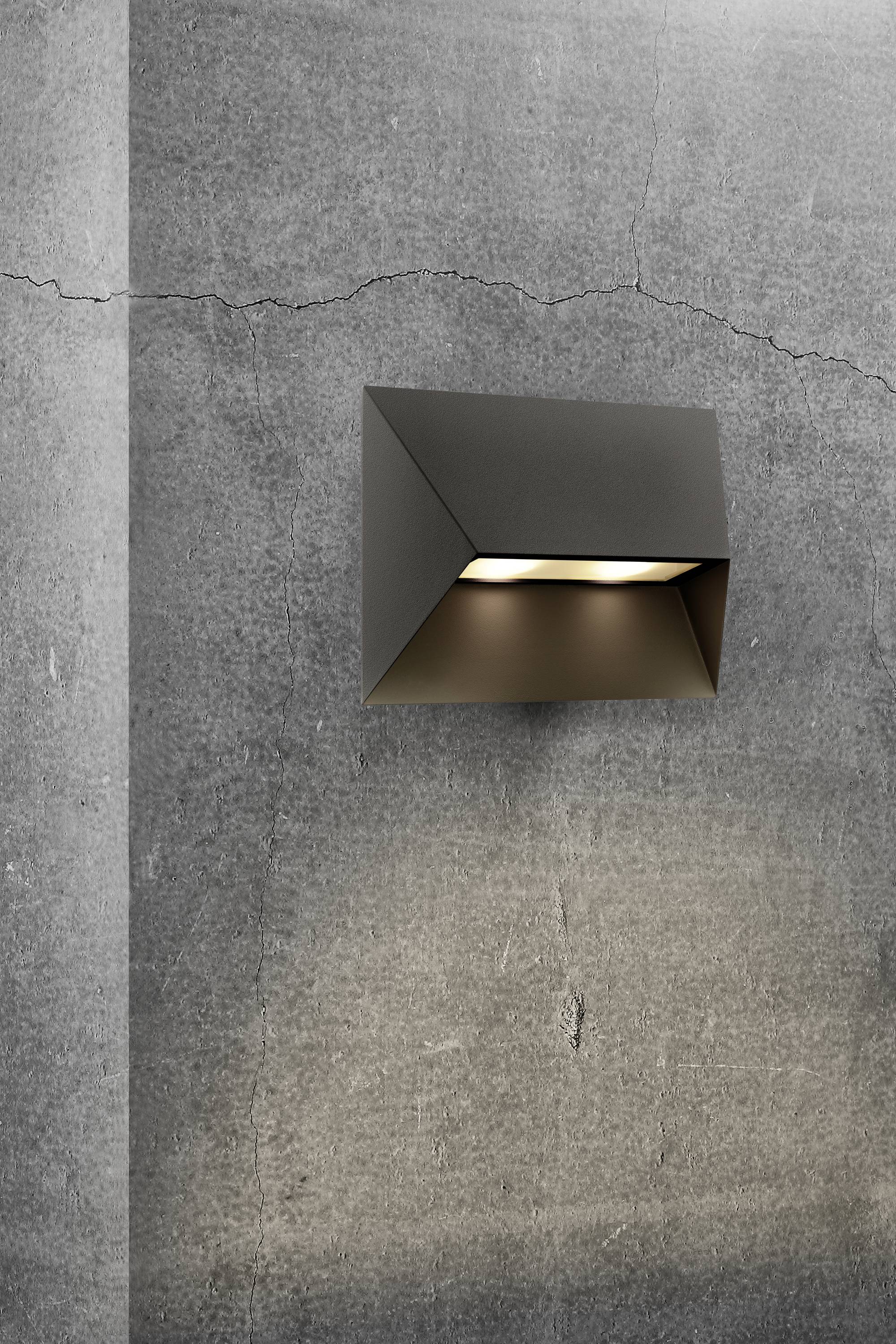 Wandleuchte aus Metall an einer Betonwand. Sie gibt warmes Licht nach unten ab und betont die raue Struktur der Wand.