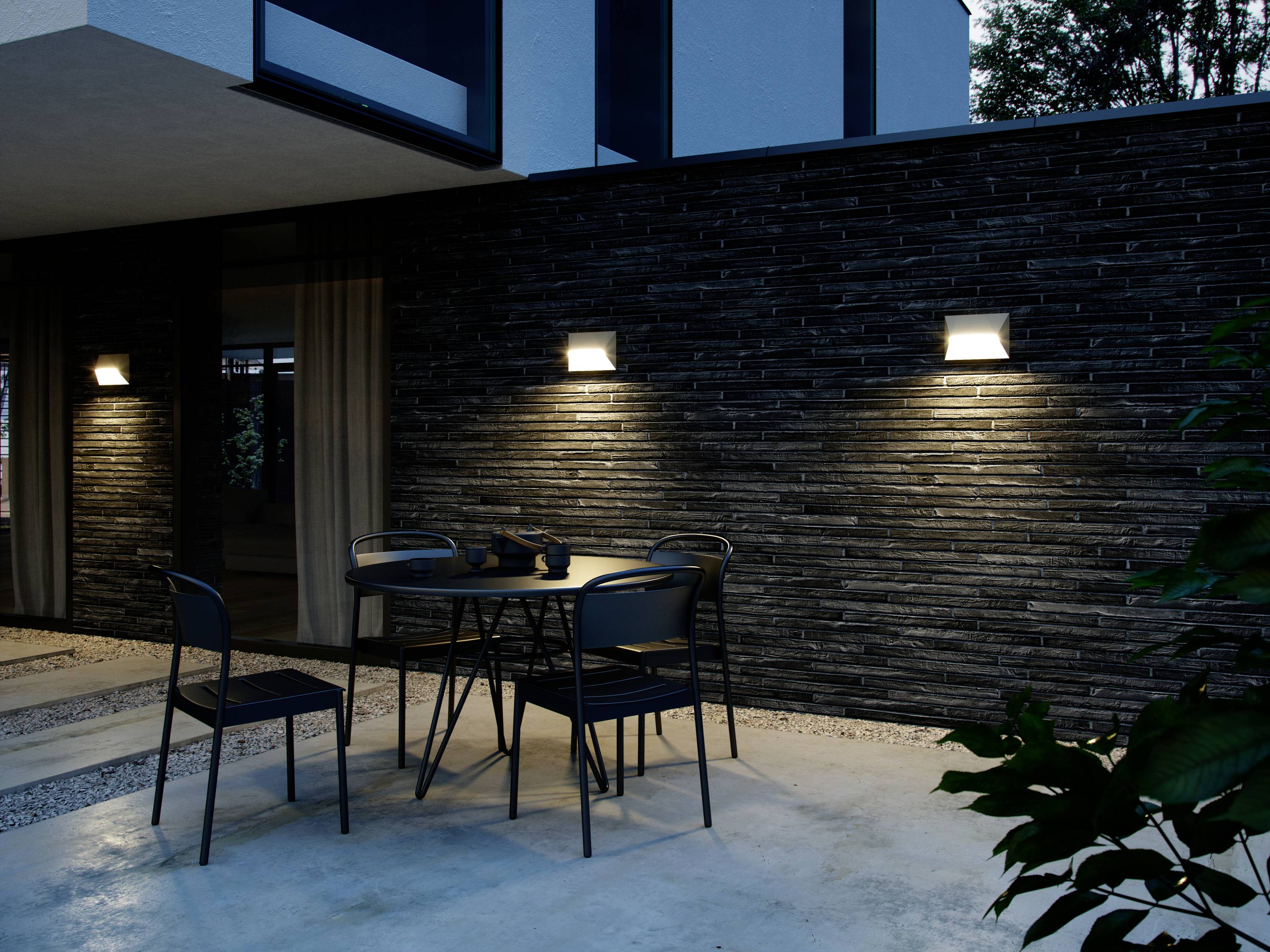 Ein moderner Außenbereich bei Abenddämmerung mit einem Tisch und Stühlen auf einer Terrasse, beleuchtet von Wandleuchten an einer dunklen Steinwand.