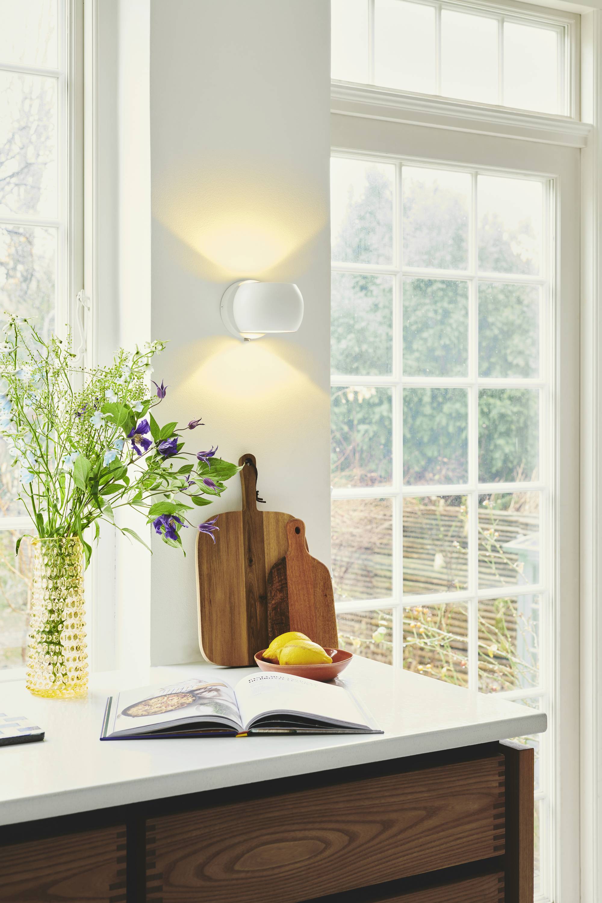 Ein heller Küchenbereich mit einem großen Fenster. Auf der Arbeitsplatte stehen Blumen in einer Vase, ein Schneidebrett und ein offenes Kochbuch.