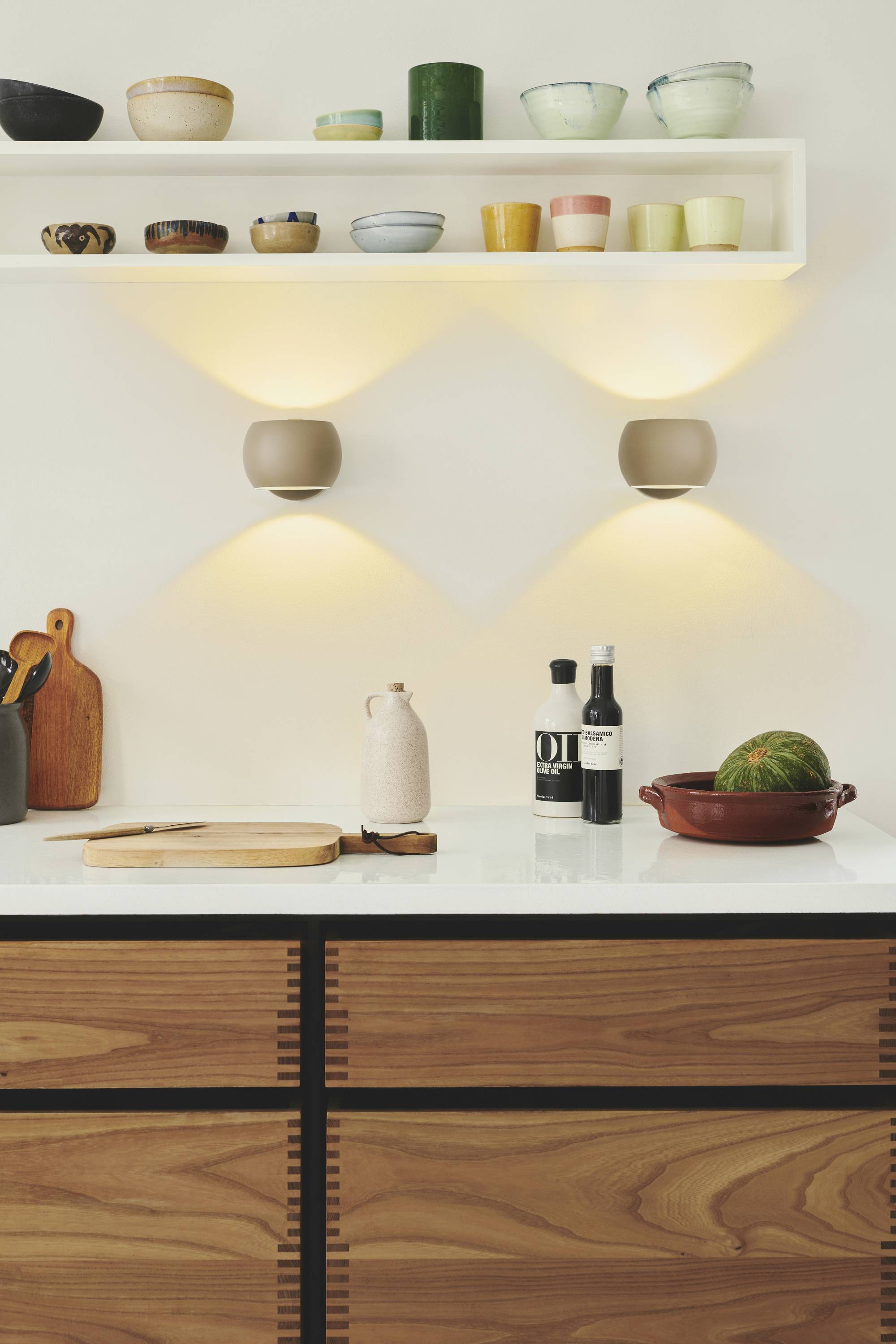 Moderne Küche mit Holzschränken, beleuchteten Wandlampen, Regal mit Keramikschalen, Brett, Gläsern und Melone auf Arbeitsplatte.