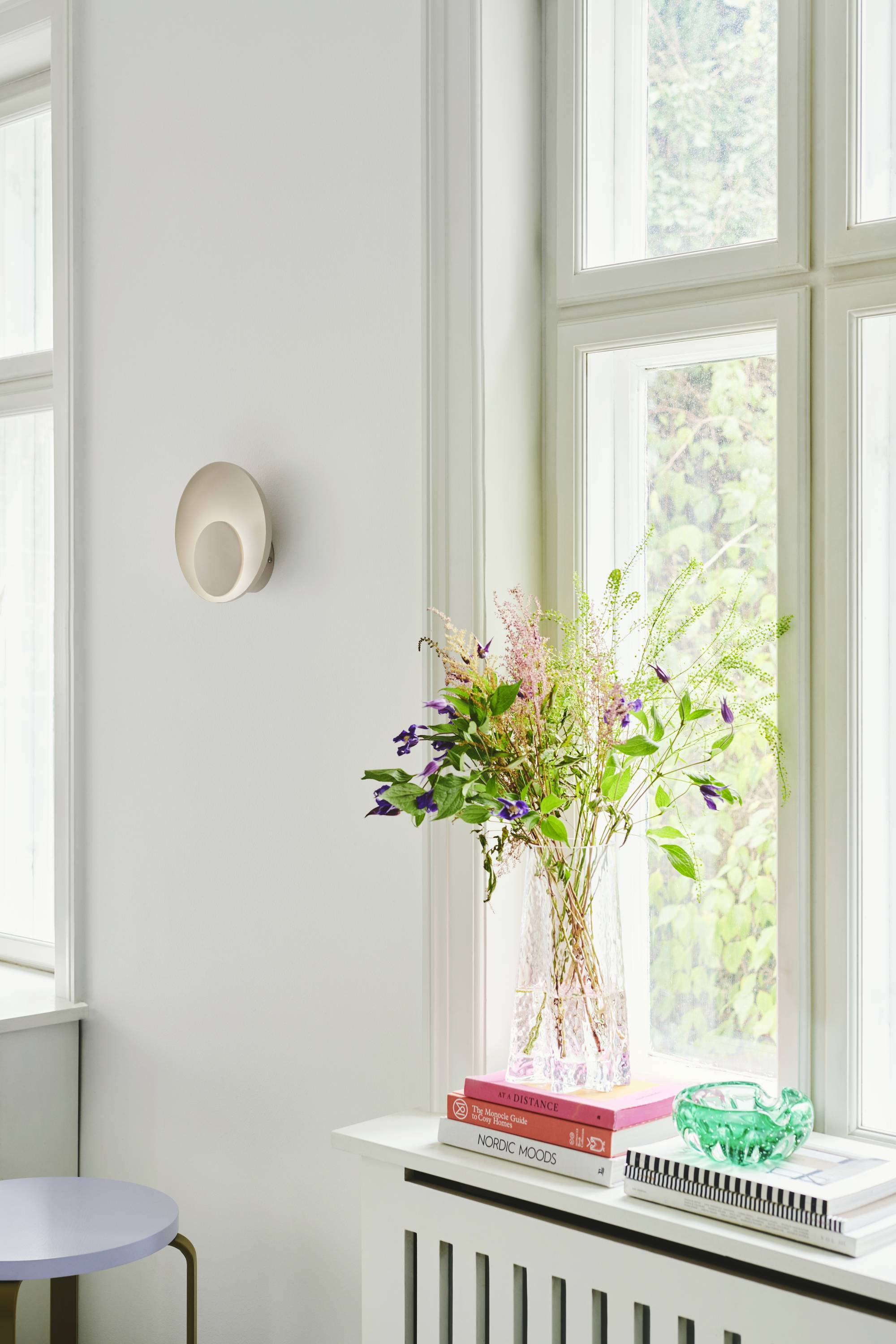 Vase mit bunten Blumen auf Fensterbank, daneben Bücher und eine grüne Glasschale. Helle, luftige Raumgestaltung mit großen Fenstern.