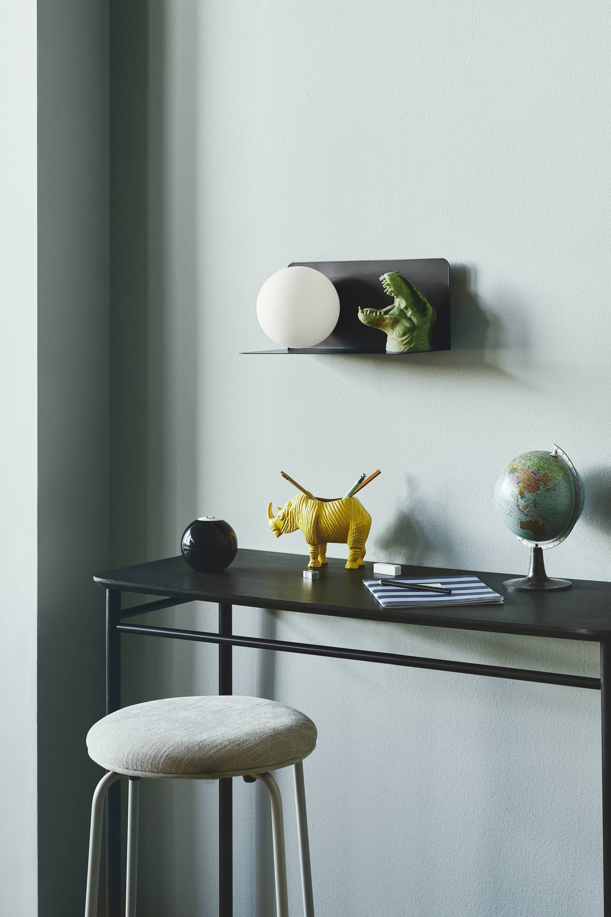 Ein schwarzer Tisch mit Hocker, Globus, gelbem Nashorn-Spielzeug und Notizbuch. Regal mit Lampe und Krokodilfigürchen darüber.