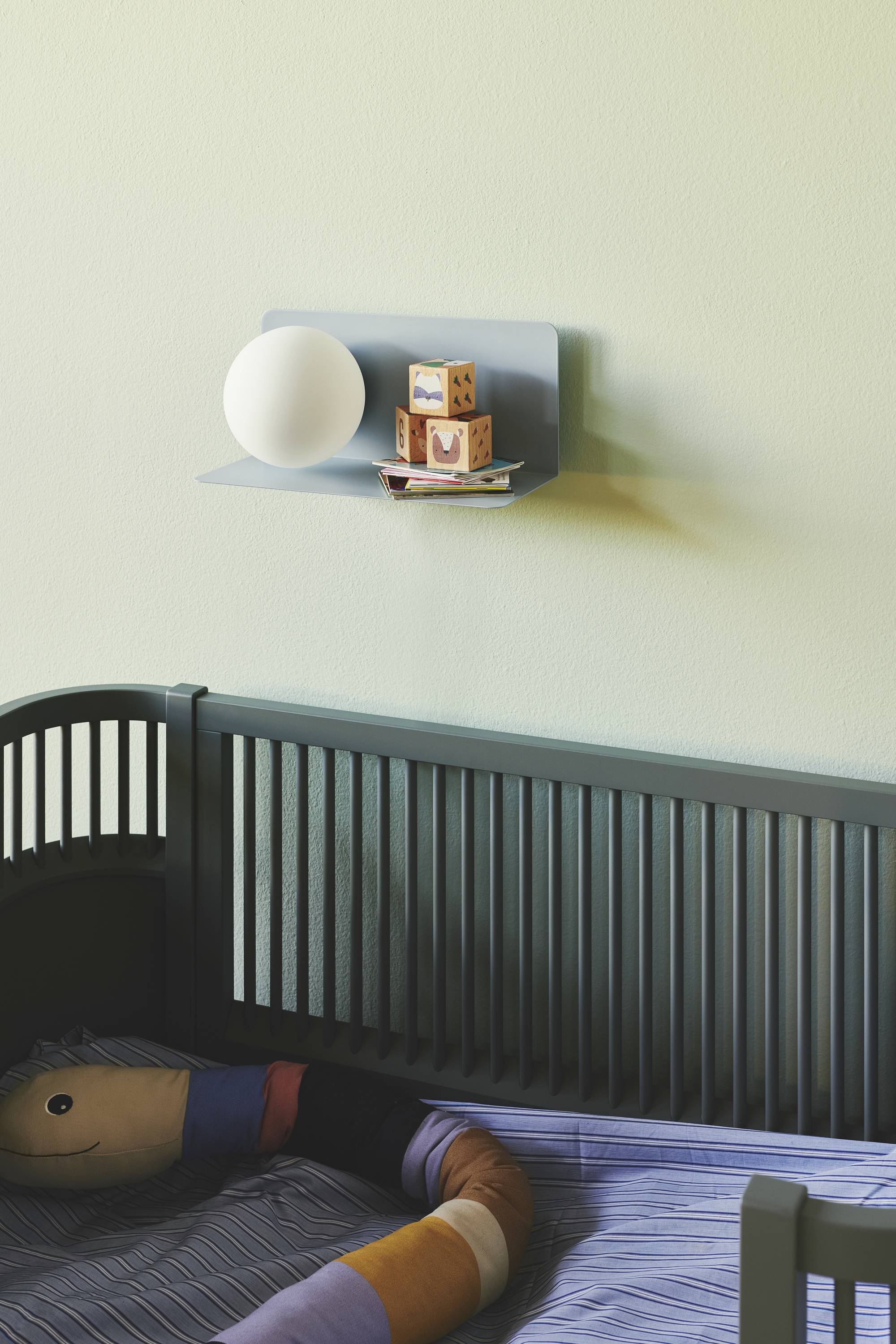 Ein Babybett mit dunklen Gitterstäben steht vor einer Wand. Auf einem Wandregal liegen eine Lampe und bunte Bausteine.