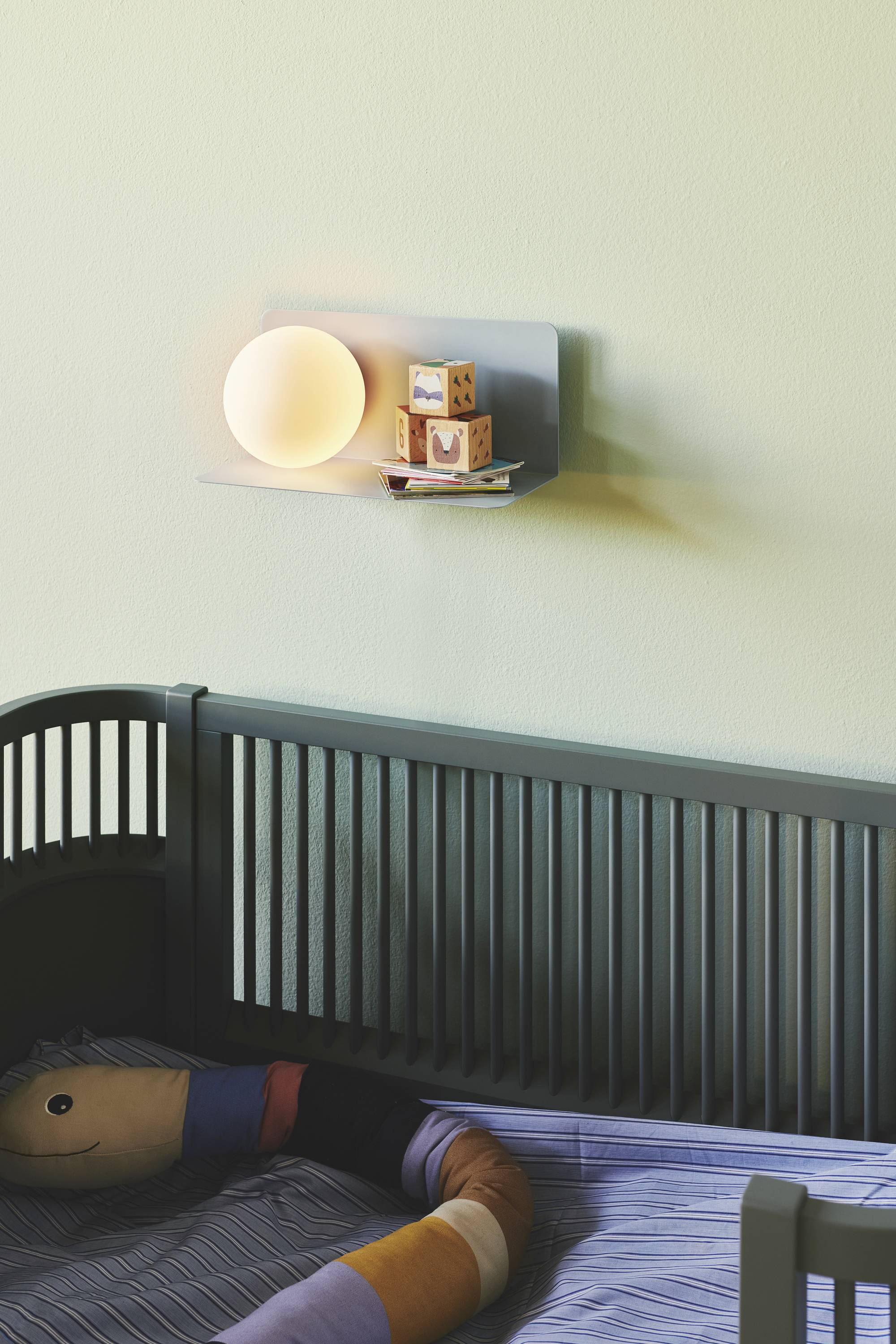 Ein Kinderbett mit dunklem Holzrahmen. Über dem Bett an der Wand ist ein Regal mit einer Lampe und Holzklötzen.
