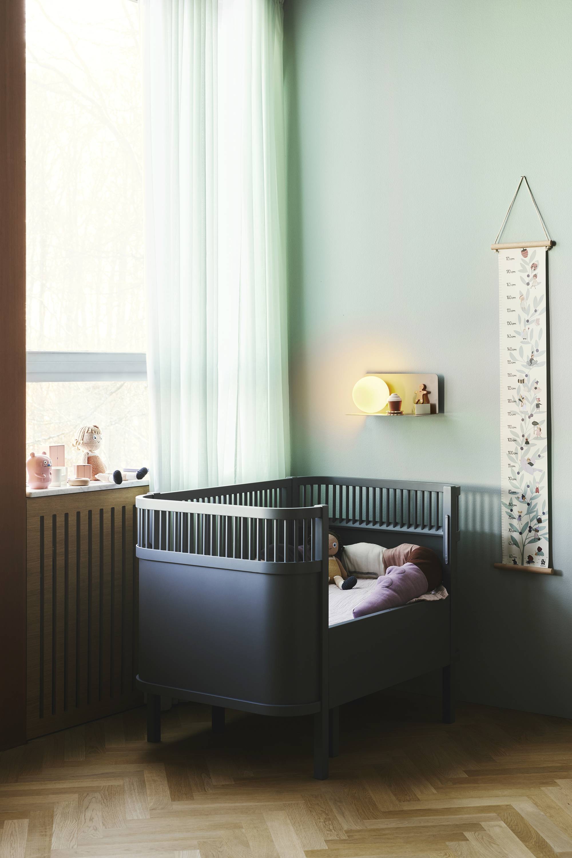 Kleines Kinderzimmer mit dunkelgrauem Kinderbett an mintgrüner Wand. Sanftes Licht von Lampe, Fenster mit fließendem Vorhang.
