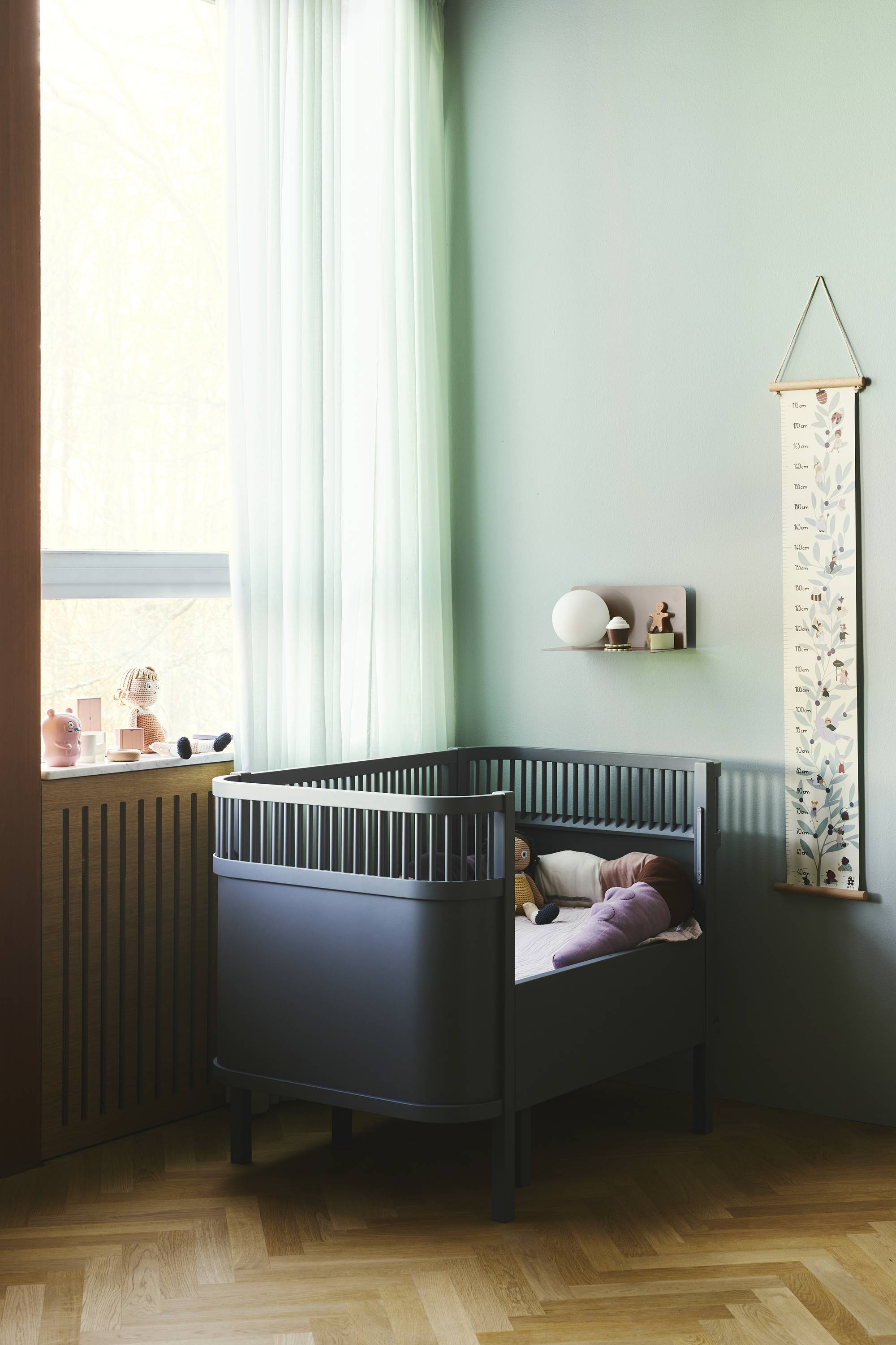 Ein Baby liegt schlafend in einem modernen, dunkelgrauen Gitterbett in einem Zimmer mit Mintgrünen Wänden und einem großen Fenster.