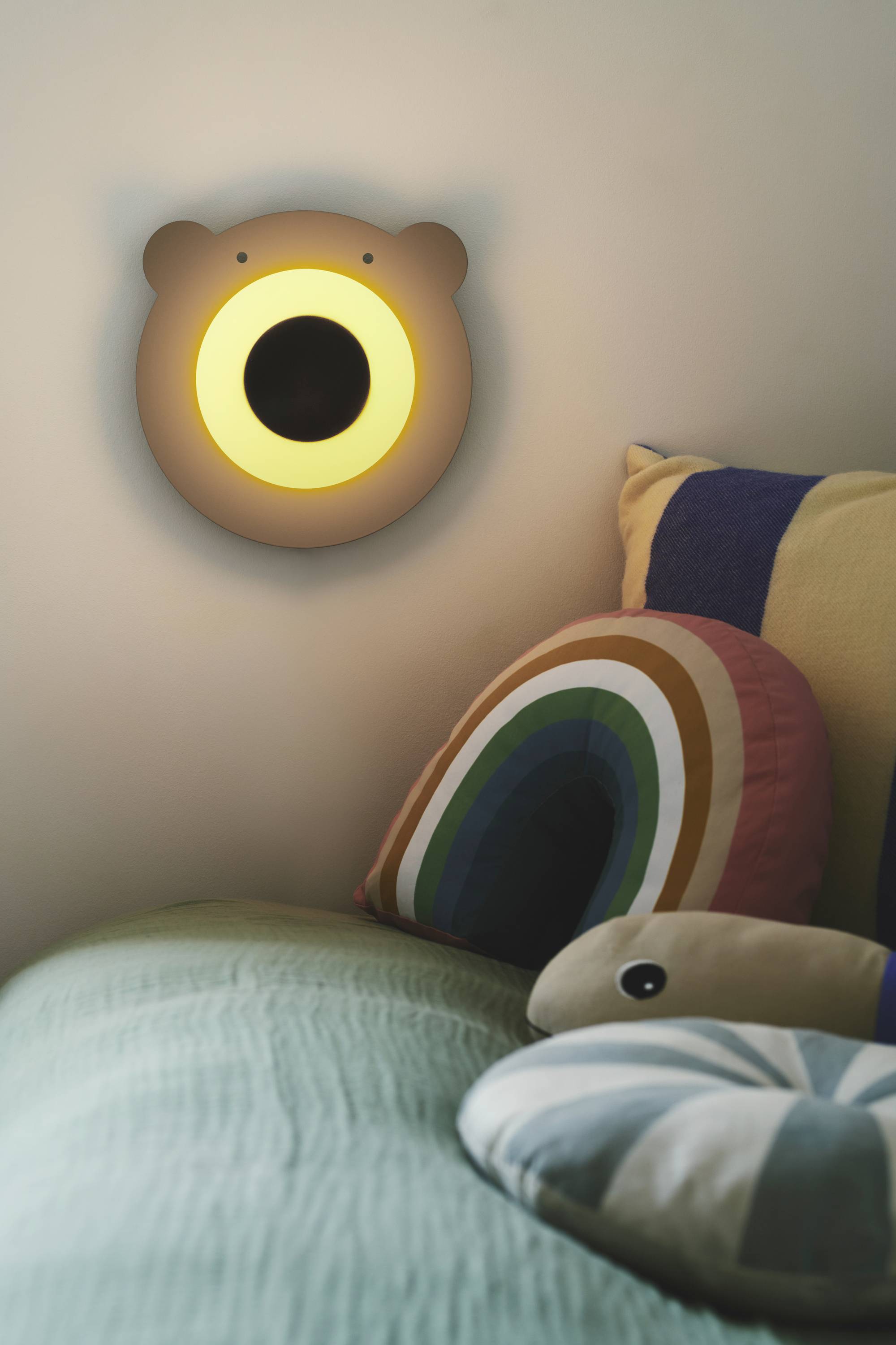 Wandlampe in Bärenform leuchtet warmes Licht über ein Sofa mit bunten Kissen, die ein Regenbogen- und Bogenmuster haben.