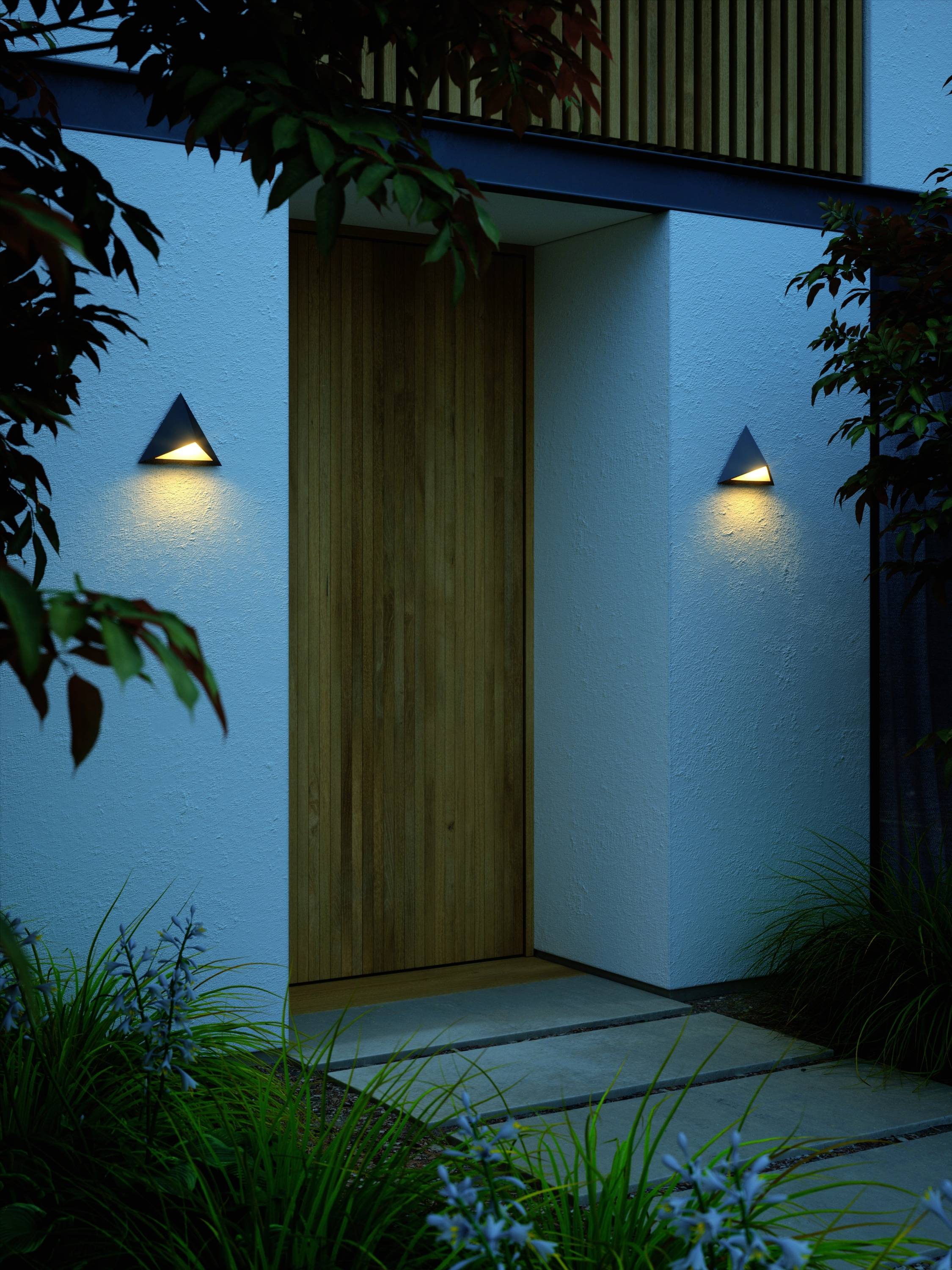 Holztür eines Hauses mit dezenten Außenleuchten, beleuchtet in der Abenddämmerung; umgeben von Pflanzen.