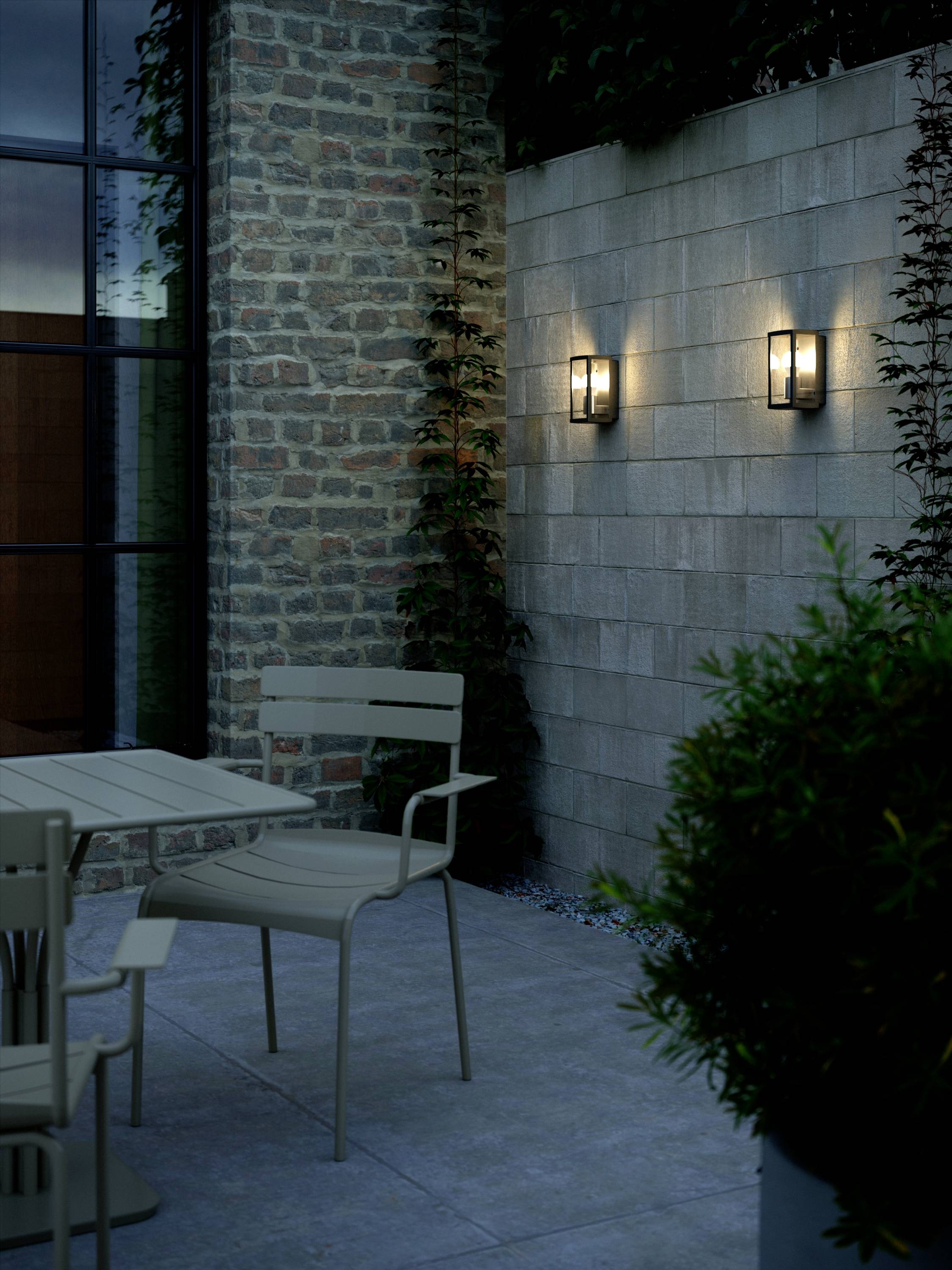 Terrasse mit Steinwänden, zwei Wandleuchten, Tisch und Stühlen. Wand mit Pflanzen bewachsen. Abendstimmung mit warmem Licht.