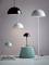 Vier moderne Lampen in verschiedenen Farben und Größen stehen auf einem Tisch und einem Podest vor einer hellgrauen Wand.