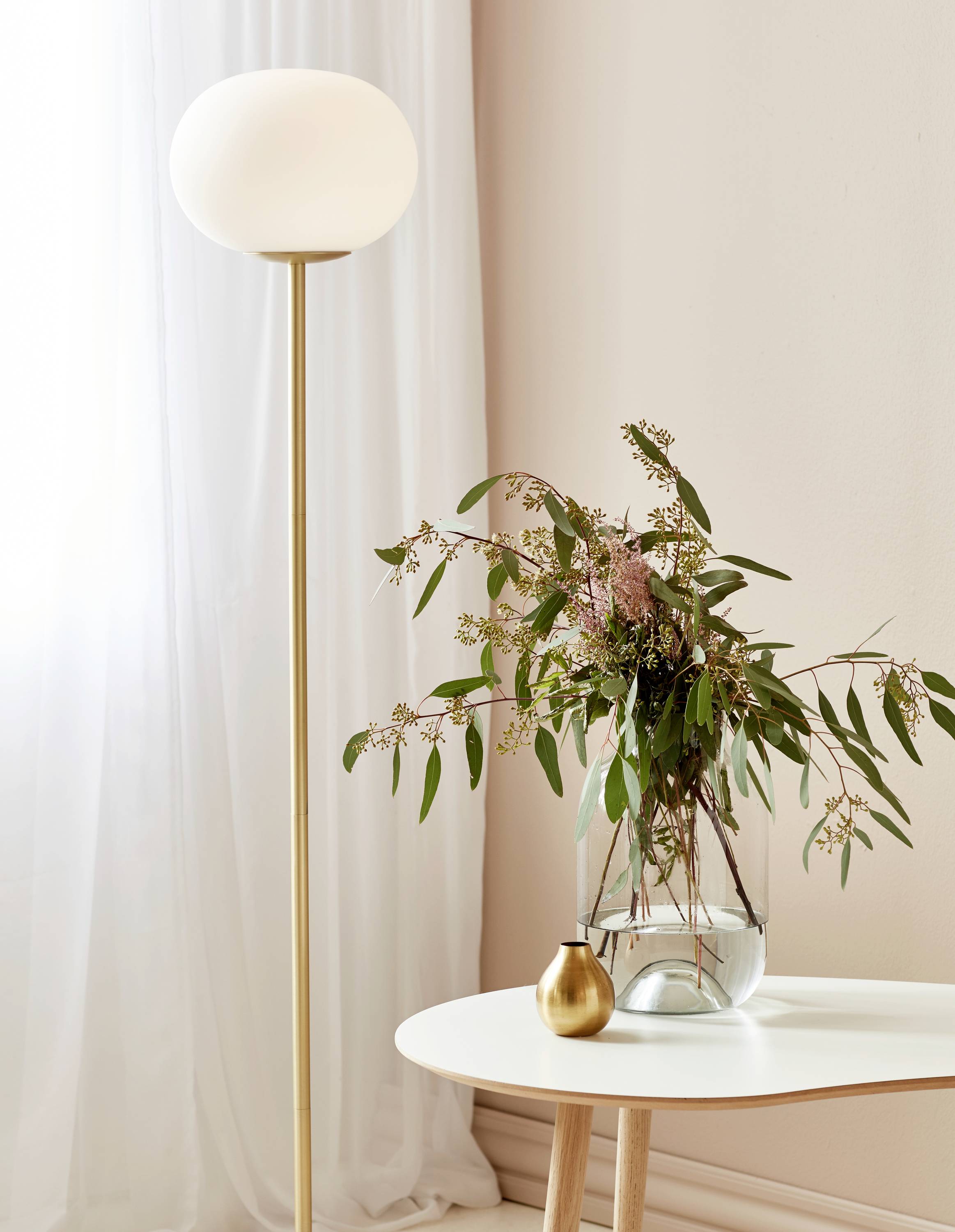 Eine minimalistische, moderne Stehlampe neben einem weißen Tisch mit einer Vase voller grüner Zweige und rosa Blumen, vor hellem Vorhang.
