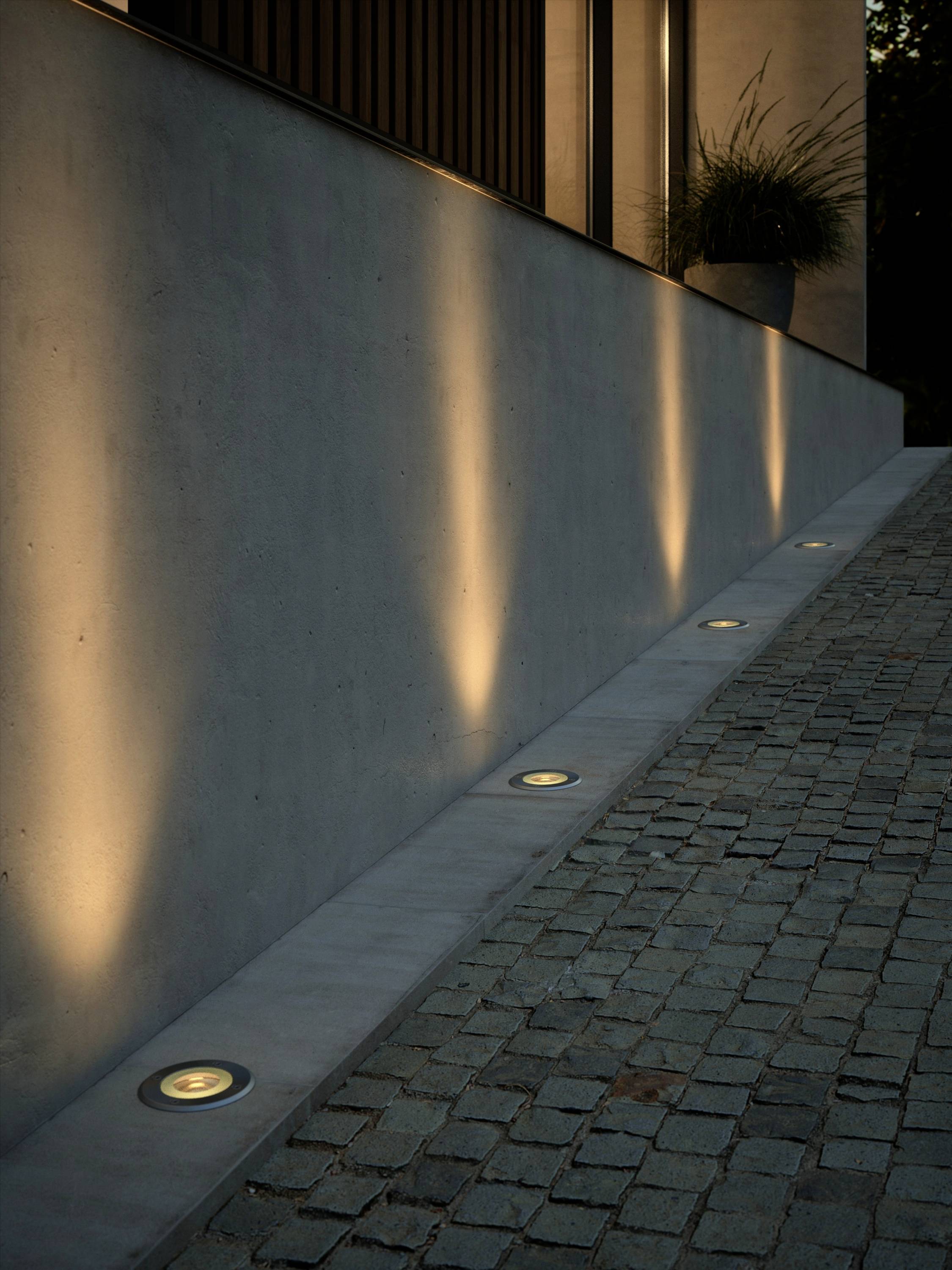 Wandleuchten beleuchten eine moderne Hausfassade und steinerne Einfahrt bei Dämmerung. Pflanzgefäß am oberen Ende der Treppe.