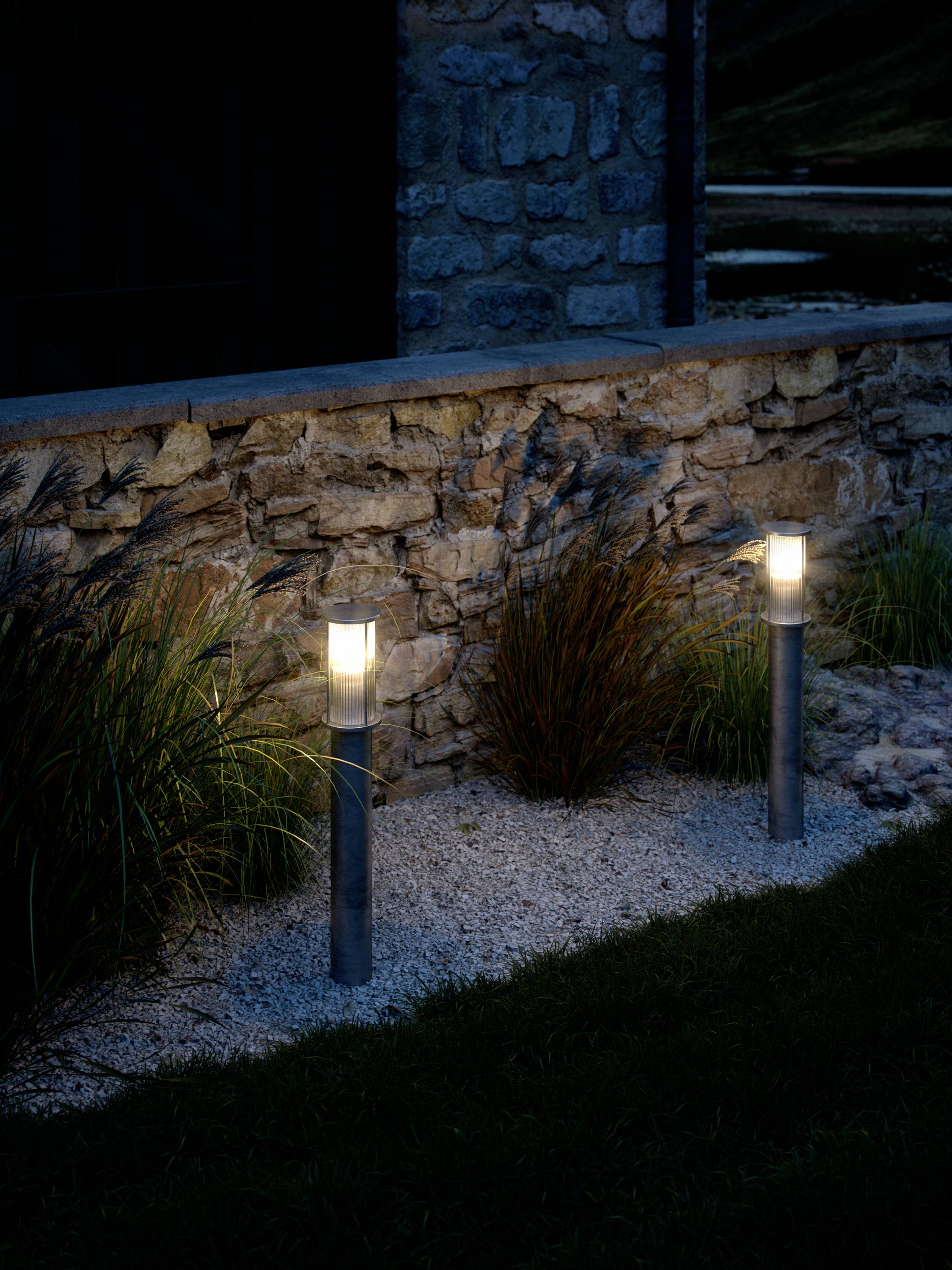 Zwei beleuchtete Gartenlampen stehen neben einer Steinmauer. Im Hintergrund ist eine grüne Wiese und ein dunkles Gebäude sichtbar.