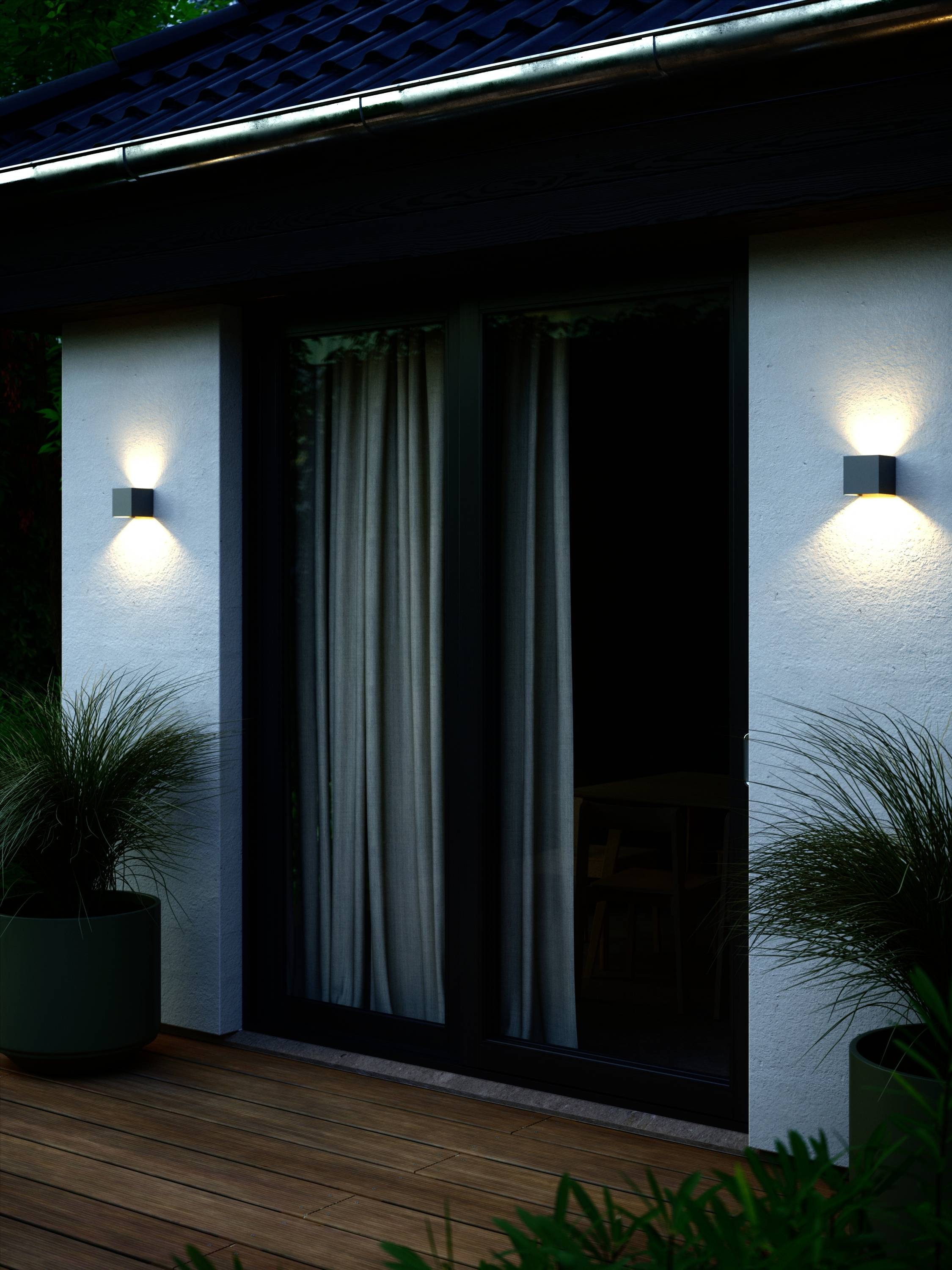 Eine moderne Terrassentür bei Nacht, daneben zwei Wandleuchten, die die Umgebung dezent beleuchten; Pflanzen in Töpfen flankieren die Tür.