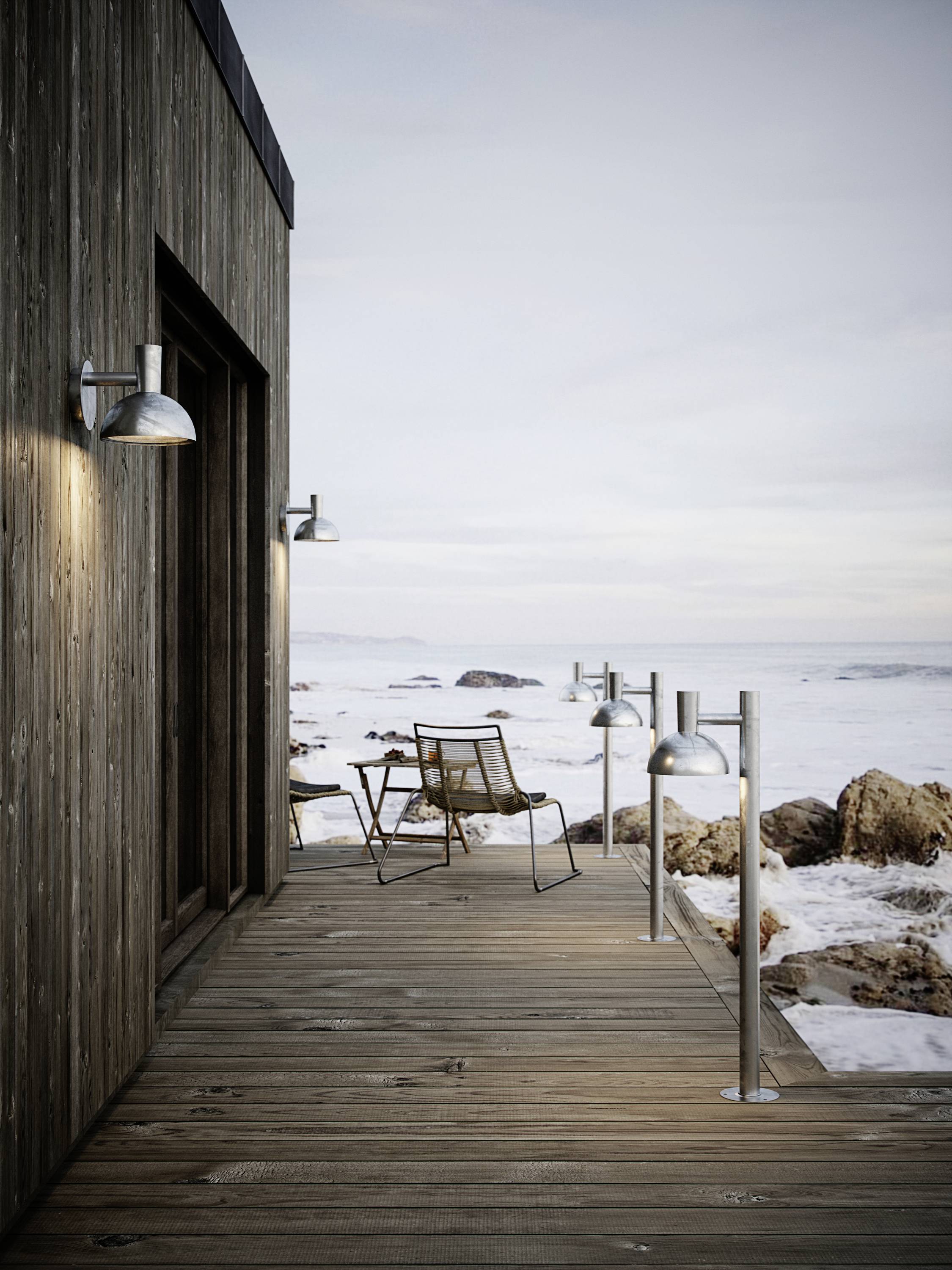 Eine Holzterrasse mit Metalllampen und zwei Stühlen, am Ufer eines felsigen Strandes. Der Himmel ist leicht bewölkt.