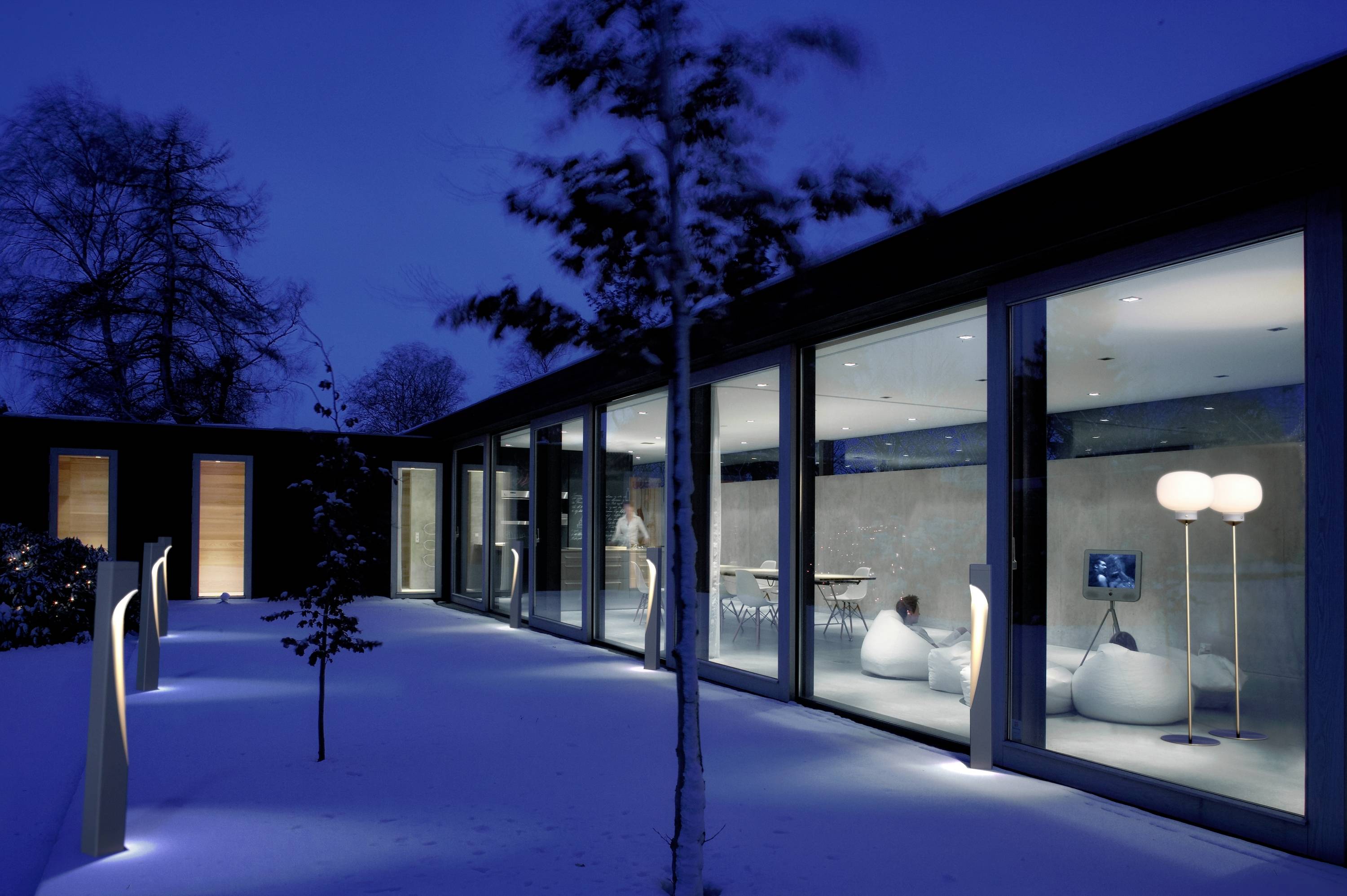 Ein modernes Gebäude mit großen Glaswänden in der Nacht. Innen sind beleuchtete Räume und Möbel sichtbar, umgeben von schneebedecktem Boden.