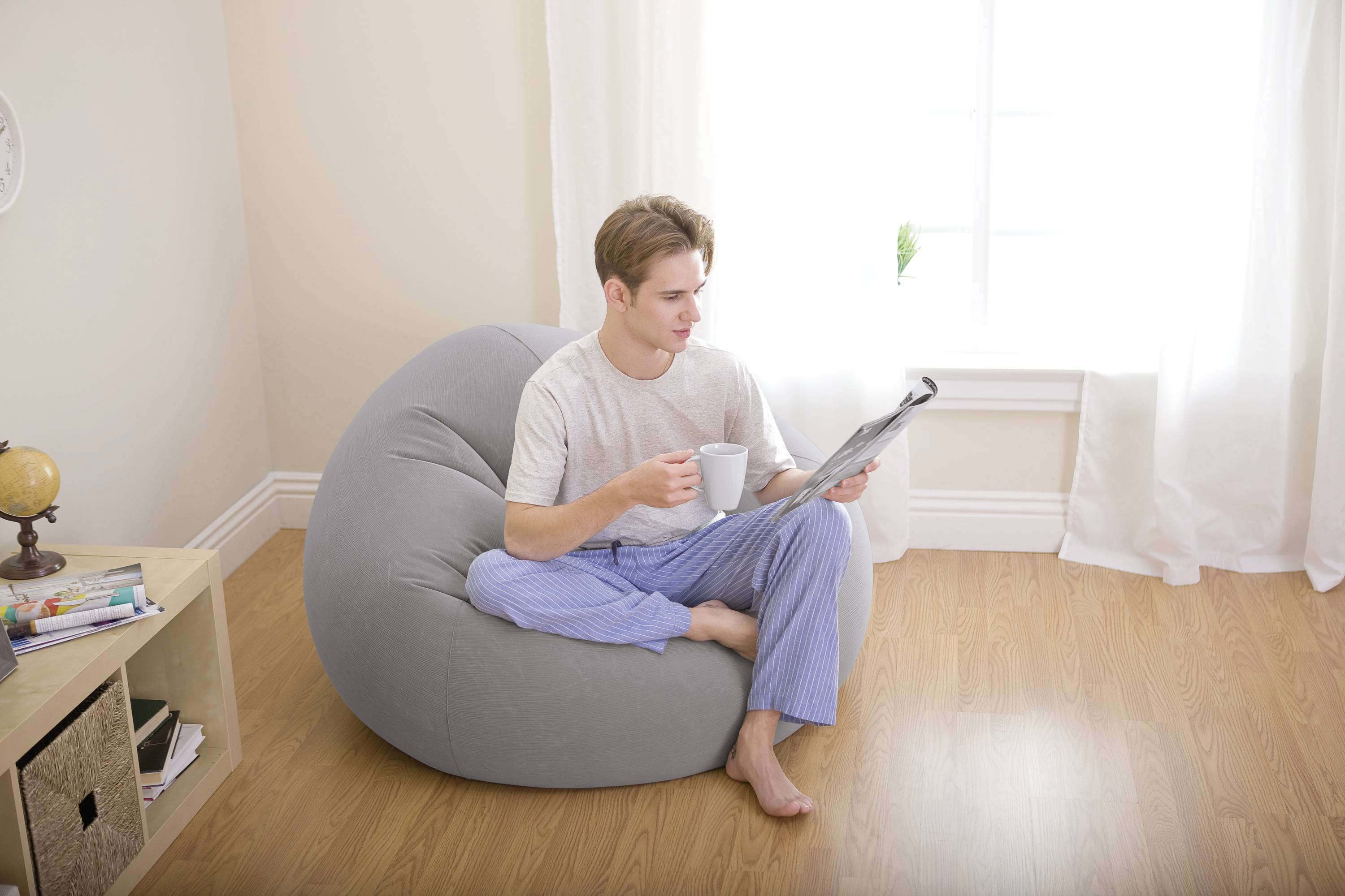 Ein Mann sitzt entspannt in einem grauen Sitzsack, hält eine Tasse und liest eine Zeitung. Er trägt ein graues T-Shirt und blaue Pyjamahosen.