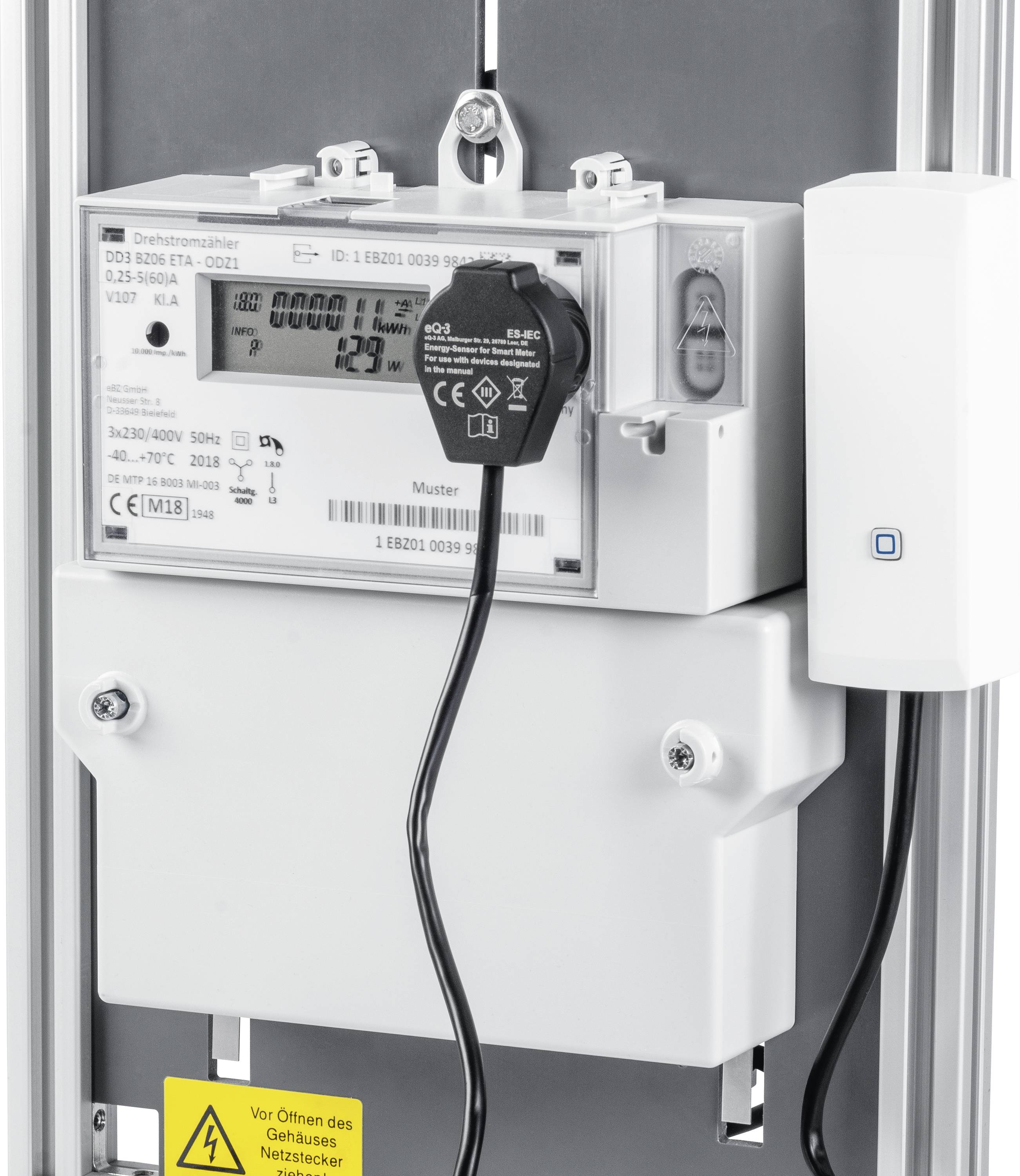 Ein digitaler Stromzähler zeigt den aktuellen Verbrauch an. Ein schwarzes Kabel ist angeschlossen. Ein Warnschild unten weist auf Vorsicht hin.