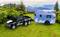 Ein schwarzes Spielzeugauto mit sechs Rädern zieht einen blauen Wohnwagen mit Bergmotiven über eine grasbewachsene Landschaft vor Bergkulisse.