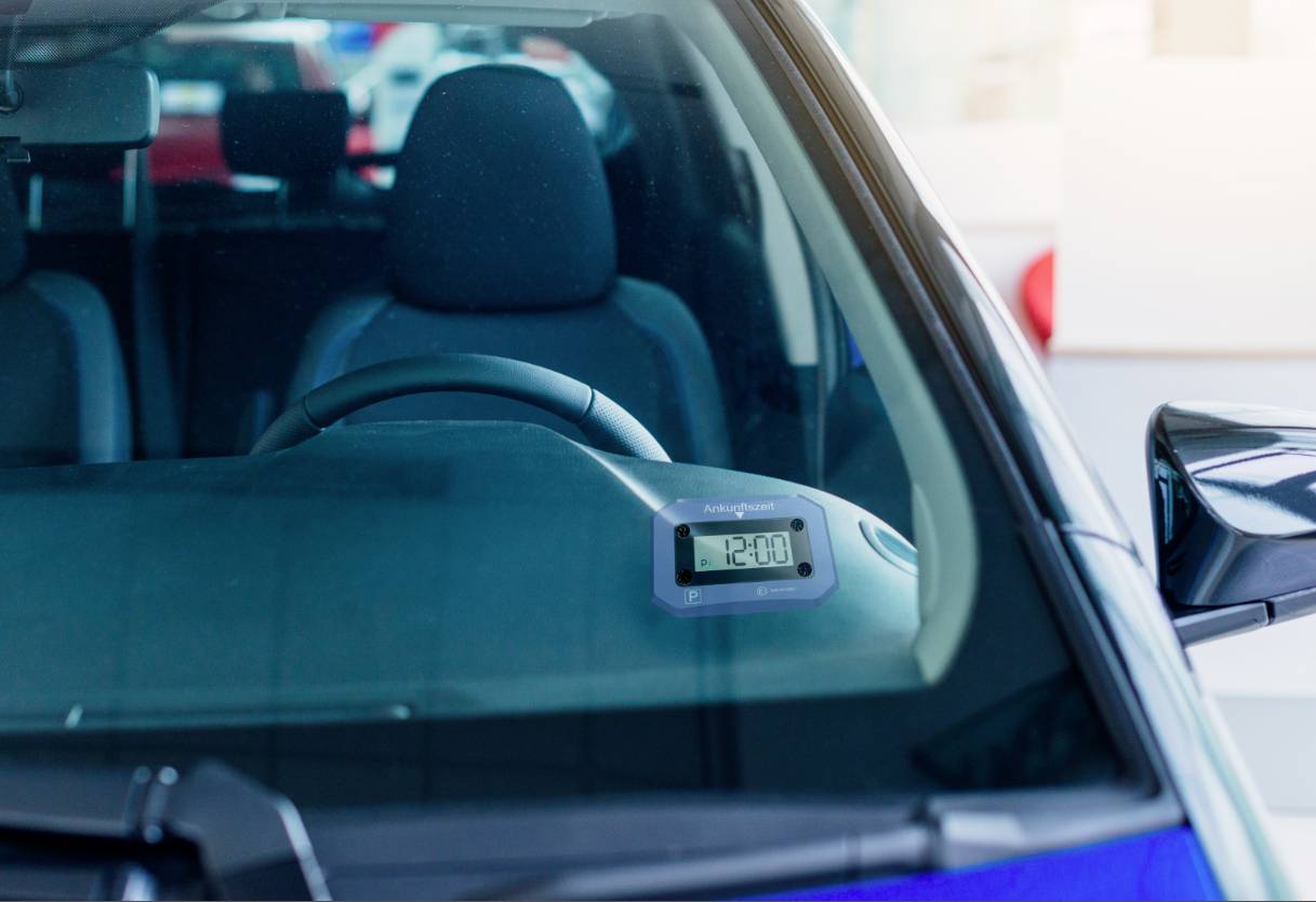 Digitale Parkscheibe im Auto zeigt '12:00' an, hinter Windschutzscheibe, leere Vorderseite des Innenraums sichtbar.