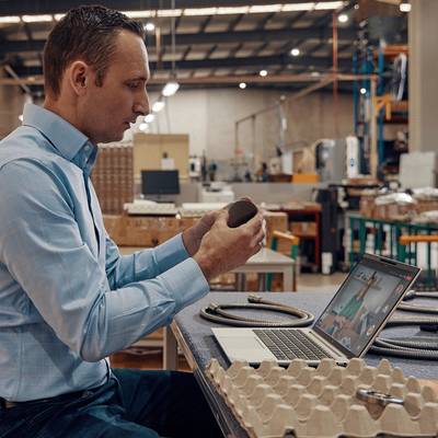 Ein Mann in einem blauen Hemd arbeitet in einer Fabrik an einem Laptop, hält ein elektronisches Bauteil und überprüft seine Eigenschaften.
