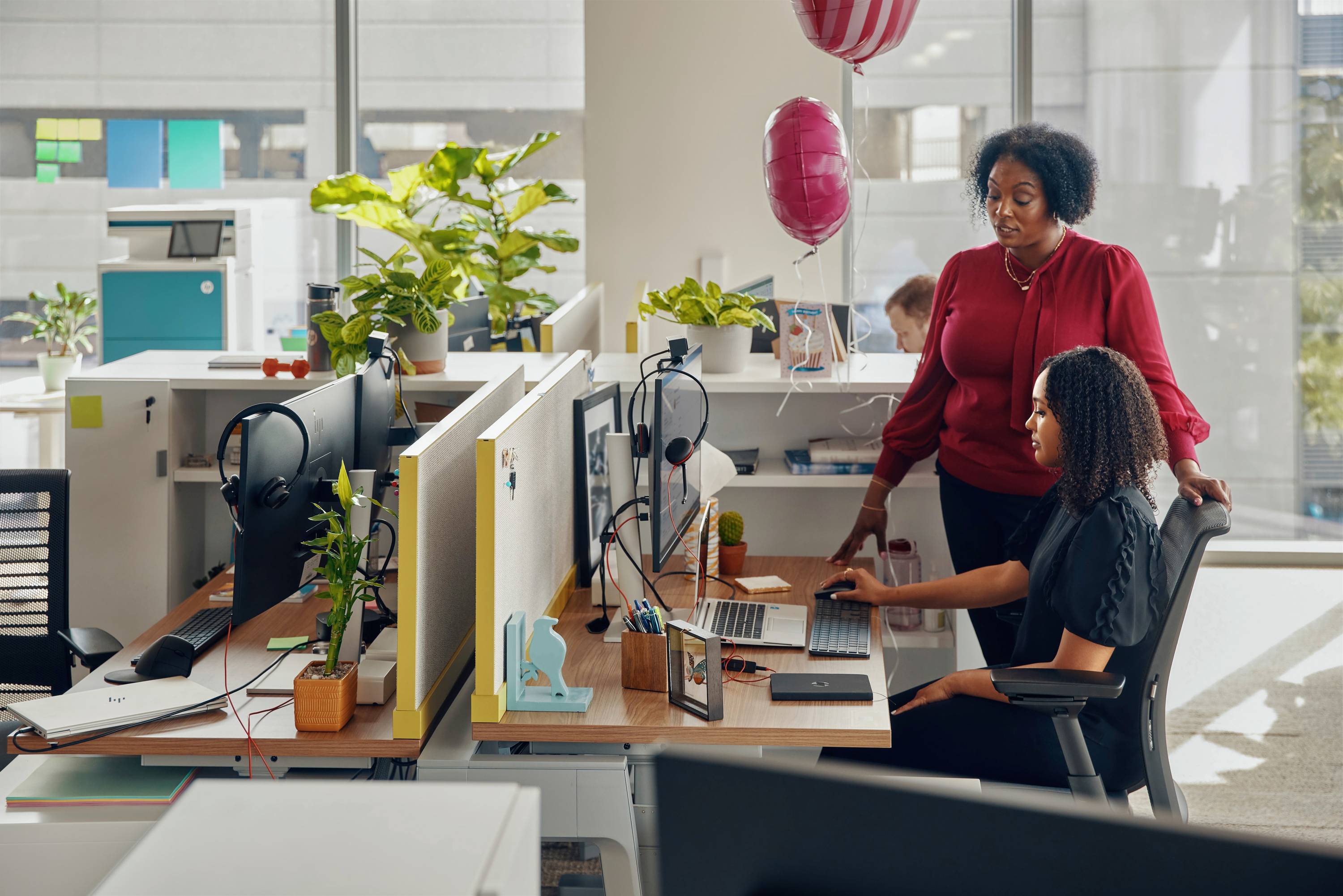 Zwei Personen arbeiten in einem modernen Büro. Eine Person sitzt am Schreibtisch, die andere steht daneben und schaut auf den Bildschirm. Luftballons und Pflanzen sind im Hintergrund.