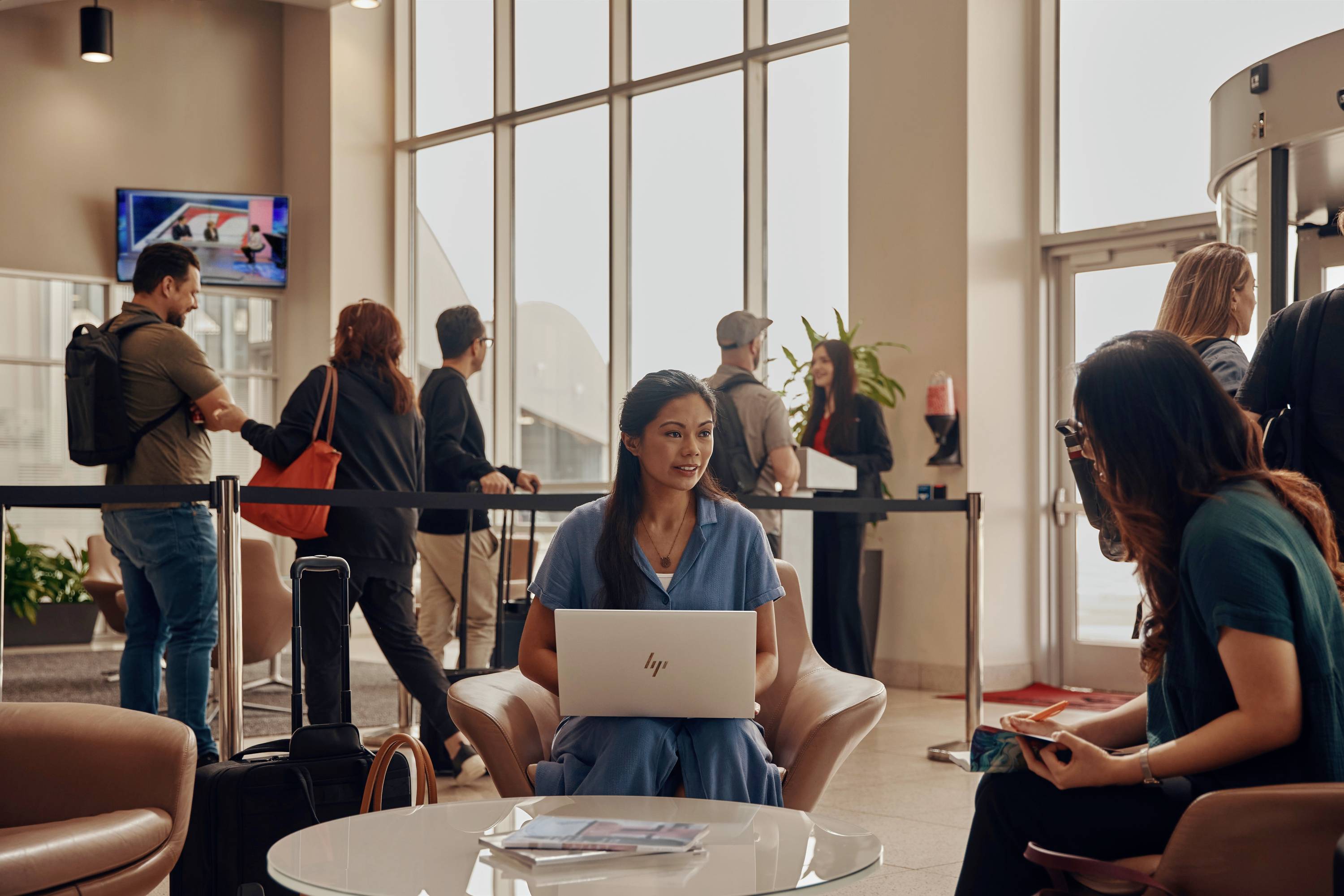 Menschen in einer belebten Flughafenlounge. Vordergrund: Zwei Frauen sitzen und unterhalten sich, eine mit einem Laptop auf dem Schoß.