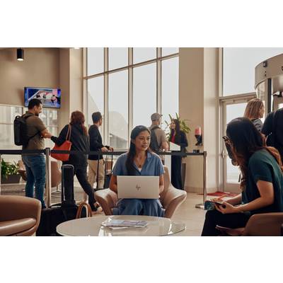 Menschen in einer belebten Flughafenlounge. Vordergrund: Zwei Frauen sitzen und unterhalten sich, eine mit einem Laptop auf dem Schoß.