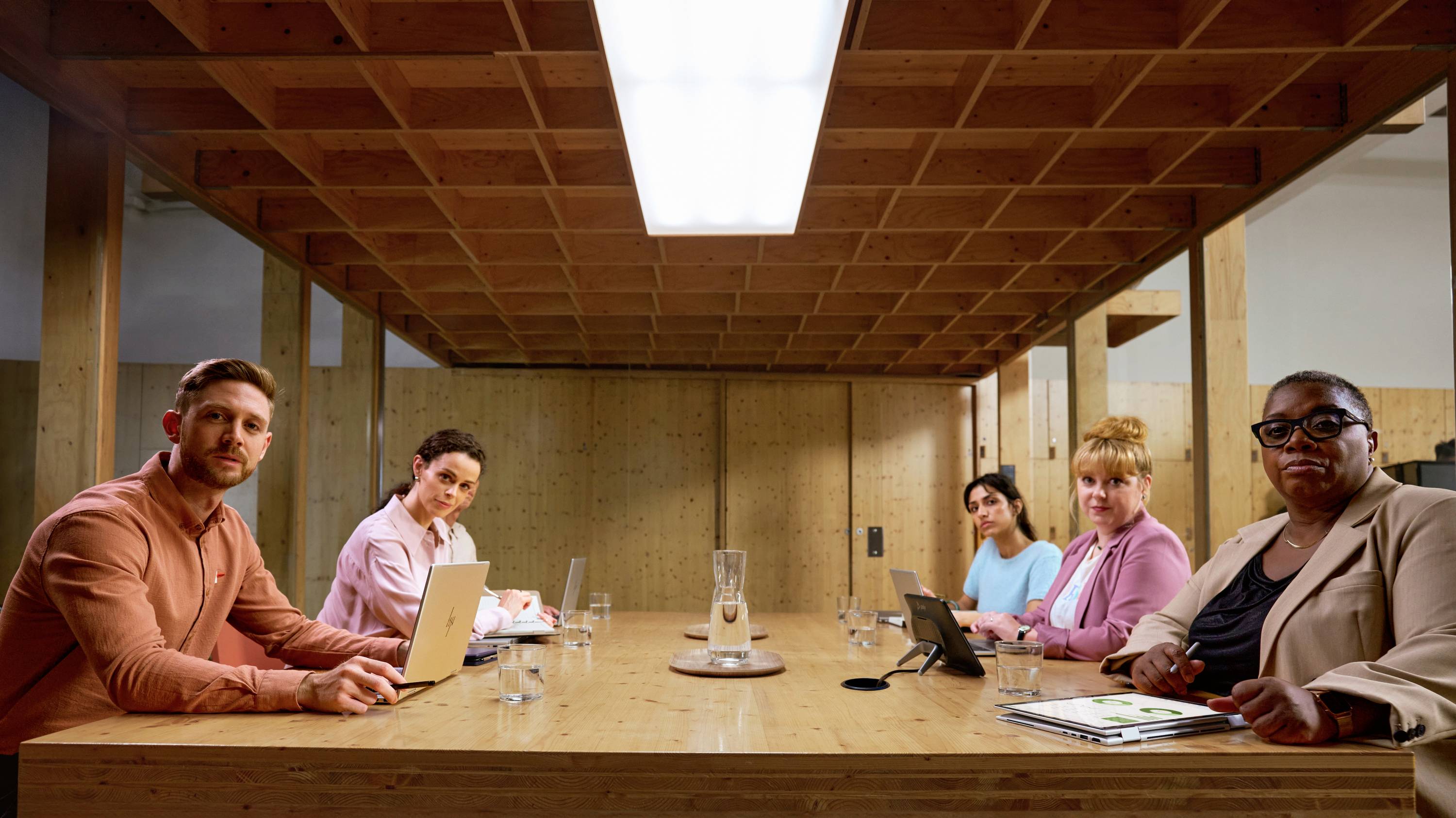 Sechs Personen sitzen an einem Konferenztisch in einem modernen Büro. Alle schauen in die Kamera, einige bedienen Laptops oder Tablets.