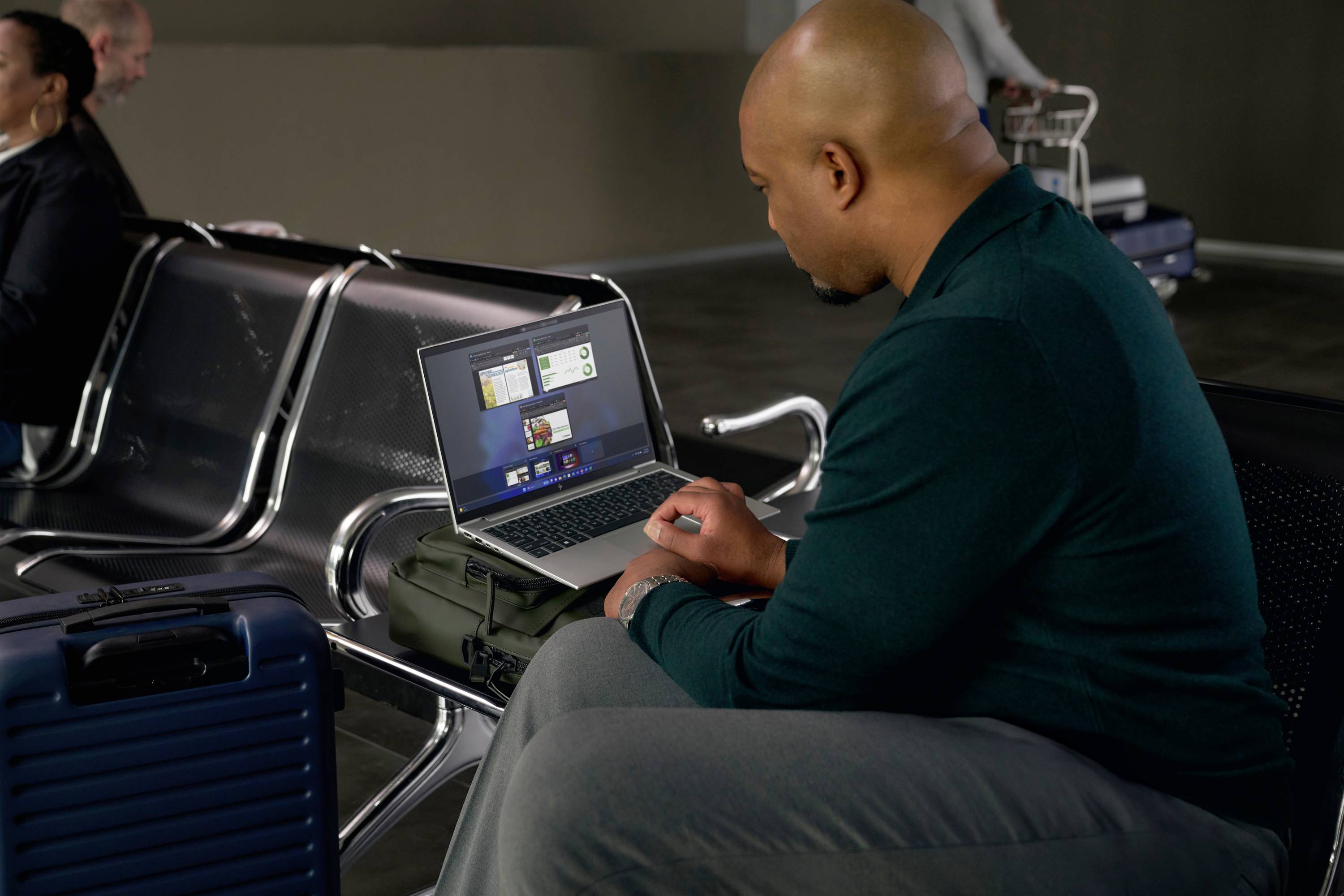 Ein Mann sitzt in einem Wartebereich am Flughafen und arbeitet auf einem Laptop, der auf seinem Handgepäck platziert ist.