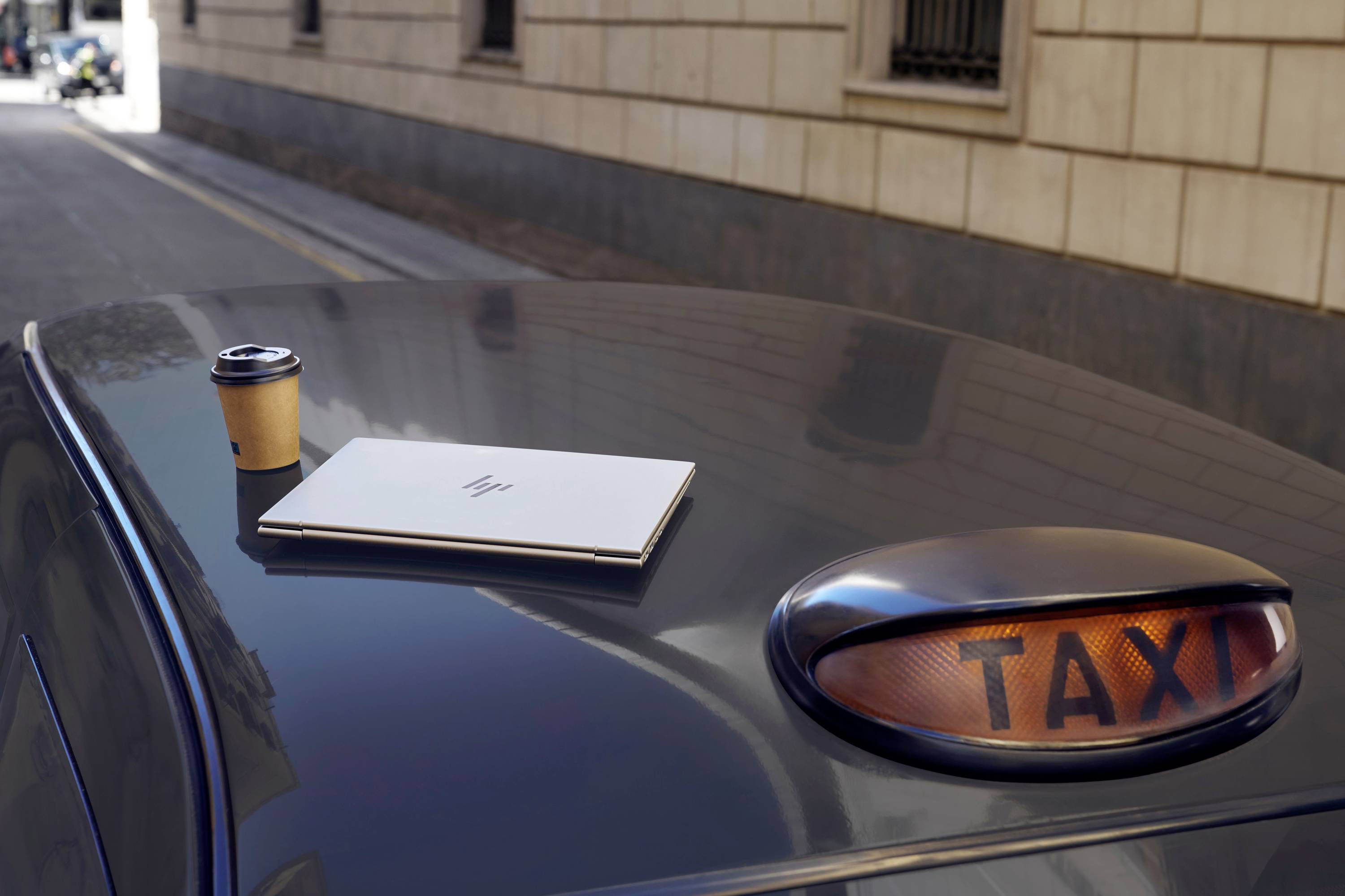 Ein Laptop und ein Kaffeebecher stehen auf dem Dach eines Taxis. Im Hintergrund sieht man eine Straße mit Gebäuden.