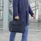 Eine Person trägt eine dunkle Tasche an der linken Hand. Sie trägt einen blauen Mantel und blaue Jeans. Hintergrund: moderne Glasarchitektur.