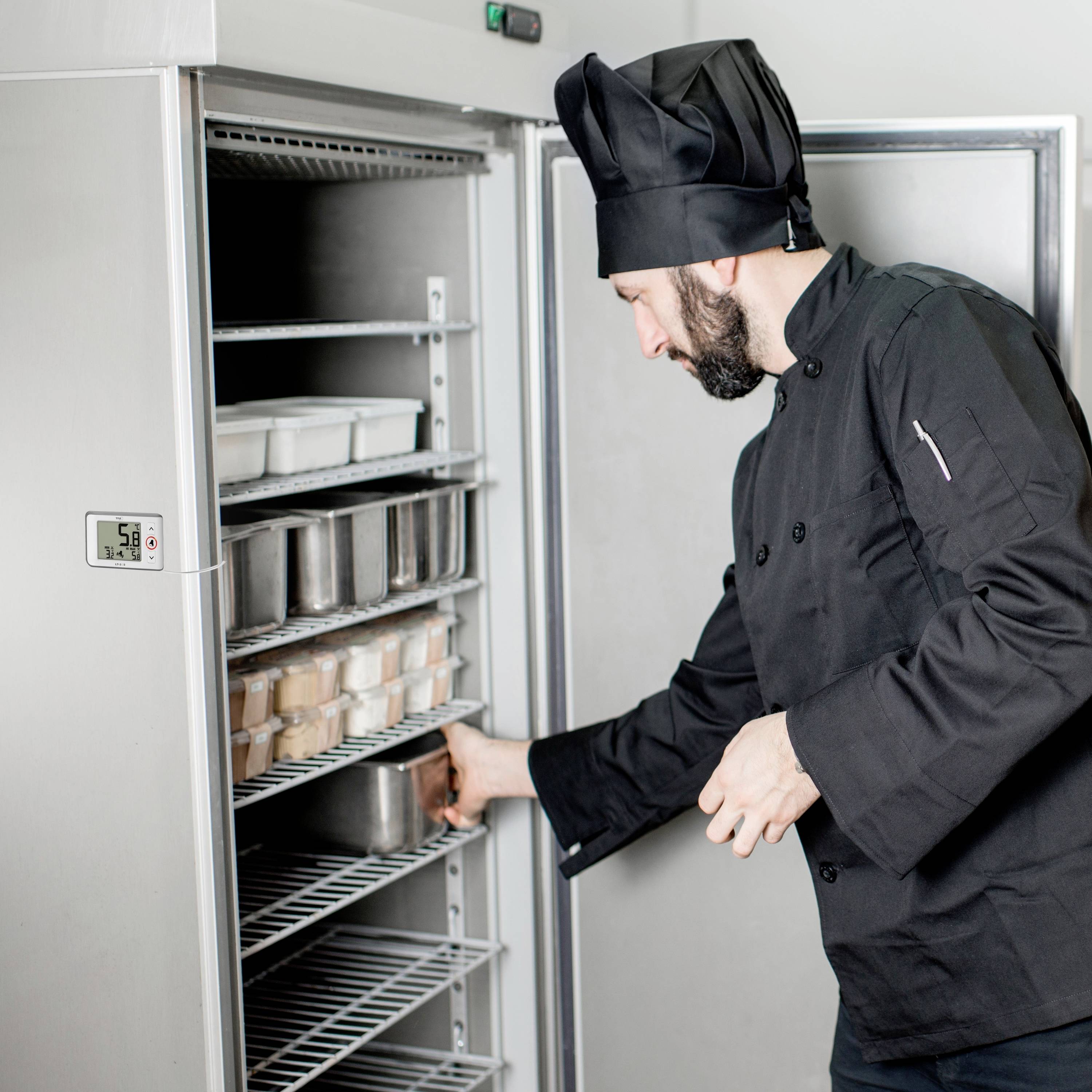 Ein Koch in schwarzer Uniform räumt Behälter in einen Edelstahl-Kühlschrank ein. Der Kühlschrank ist offen und gefüllt mit Lebensmitteln.