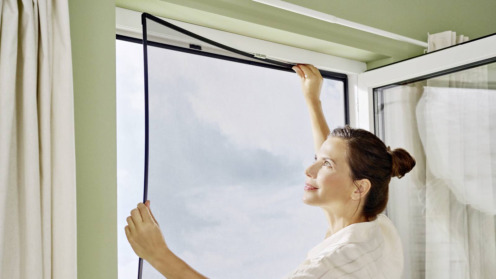 Eine Person befestigt ein schwarzes Fliegengitter an einem geöffneten Fenster, durch das blauer Himmel mit Wolken zu sehen ist.