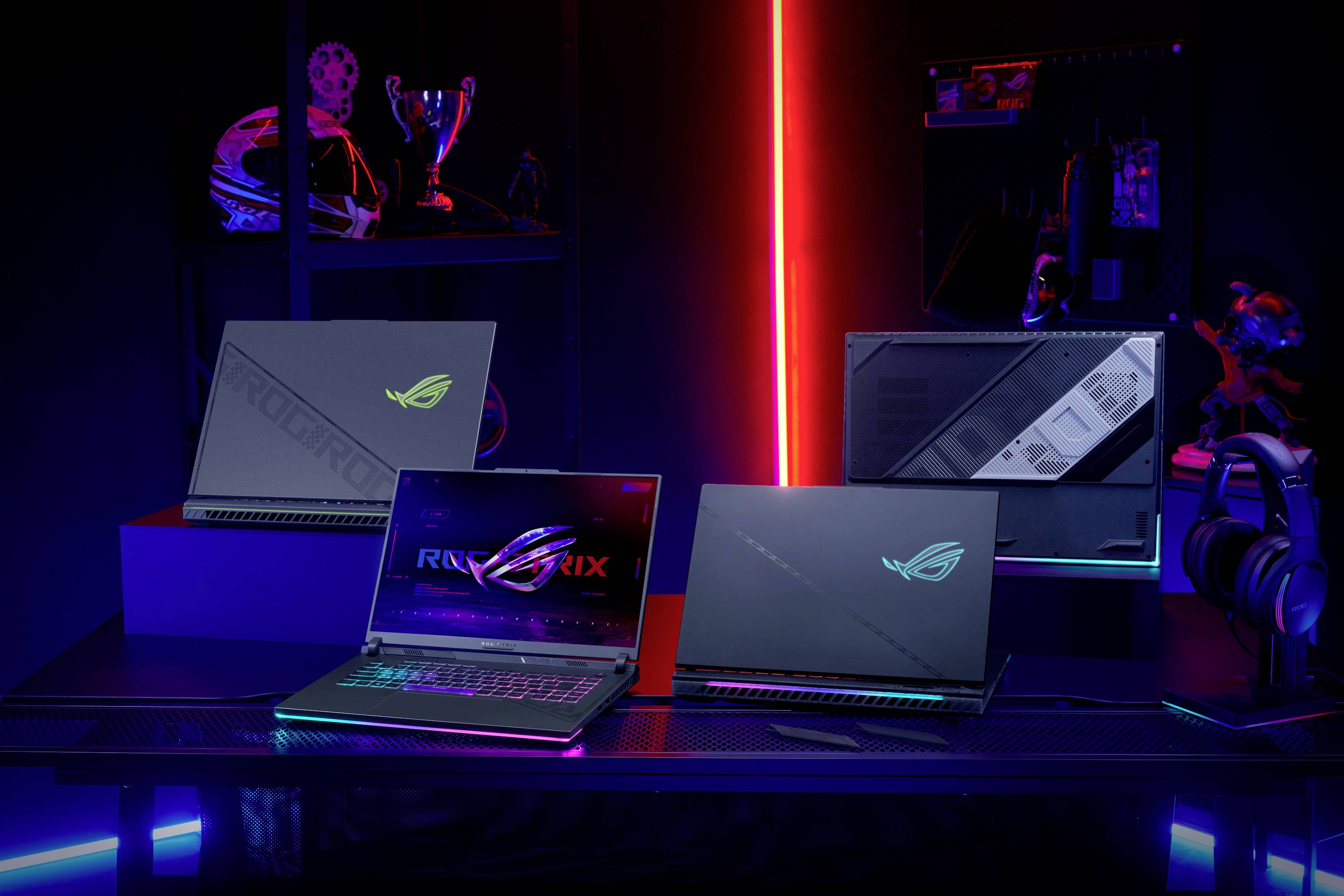 Drei Laptops auf einem Tisch in einem beleuchteten Raum. Im Hintergrund sind ein Helm, ein Pokal und Computerzubehör zu sehen.