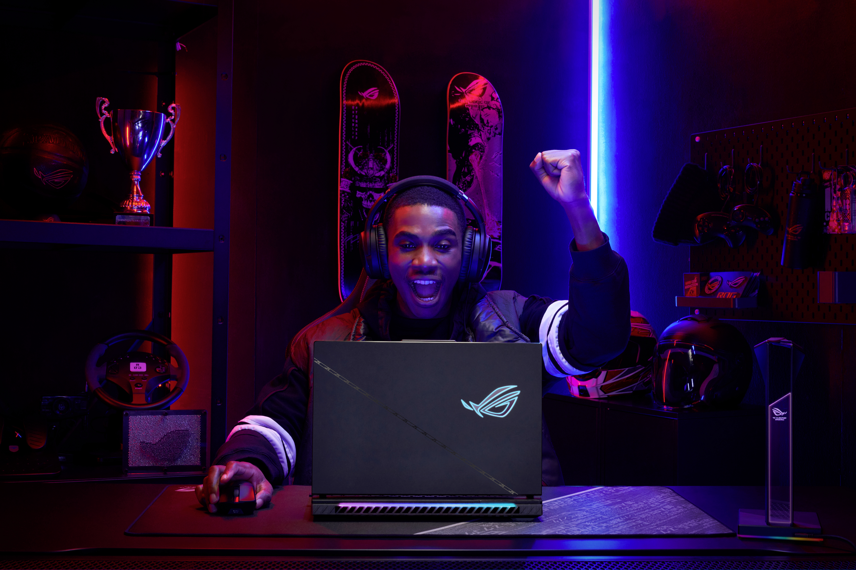 Eine Person sitzt an einem Laptop in einem Gaming-Zimmer. Sie trägt Kopfhörer und hebt triumphierend eine Faust. Im Hintergrund sind Trophäen und Gaming-Ausrüstung zu sehen.