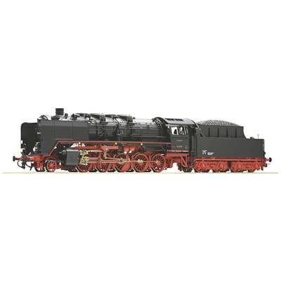 Eine schwarze Dampflokomotive mit roten Rädern und einem Kohletender wird von einem Lokführer gefahren. Sie steht auf einer Schiene.