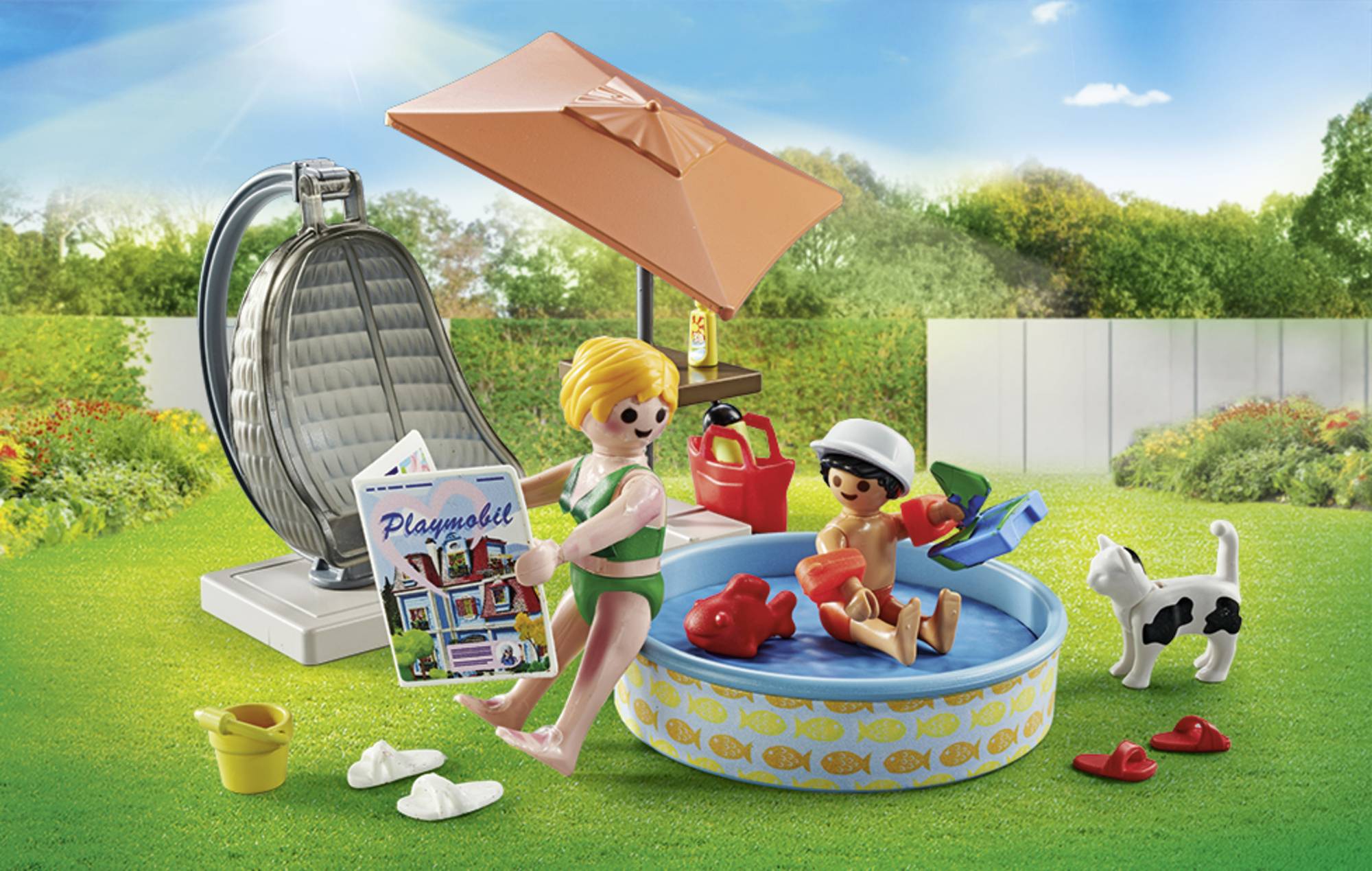 Zwei Kinder spielen im Mini-Pool im Garten. Ein Kind liest ein 'Playmobil'-Katalog. Im Hintergrund Sonnenschirm, Grill und Hund.