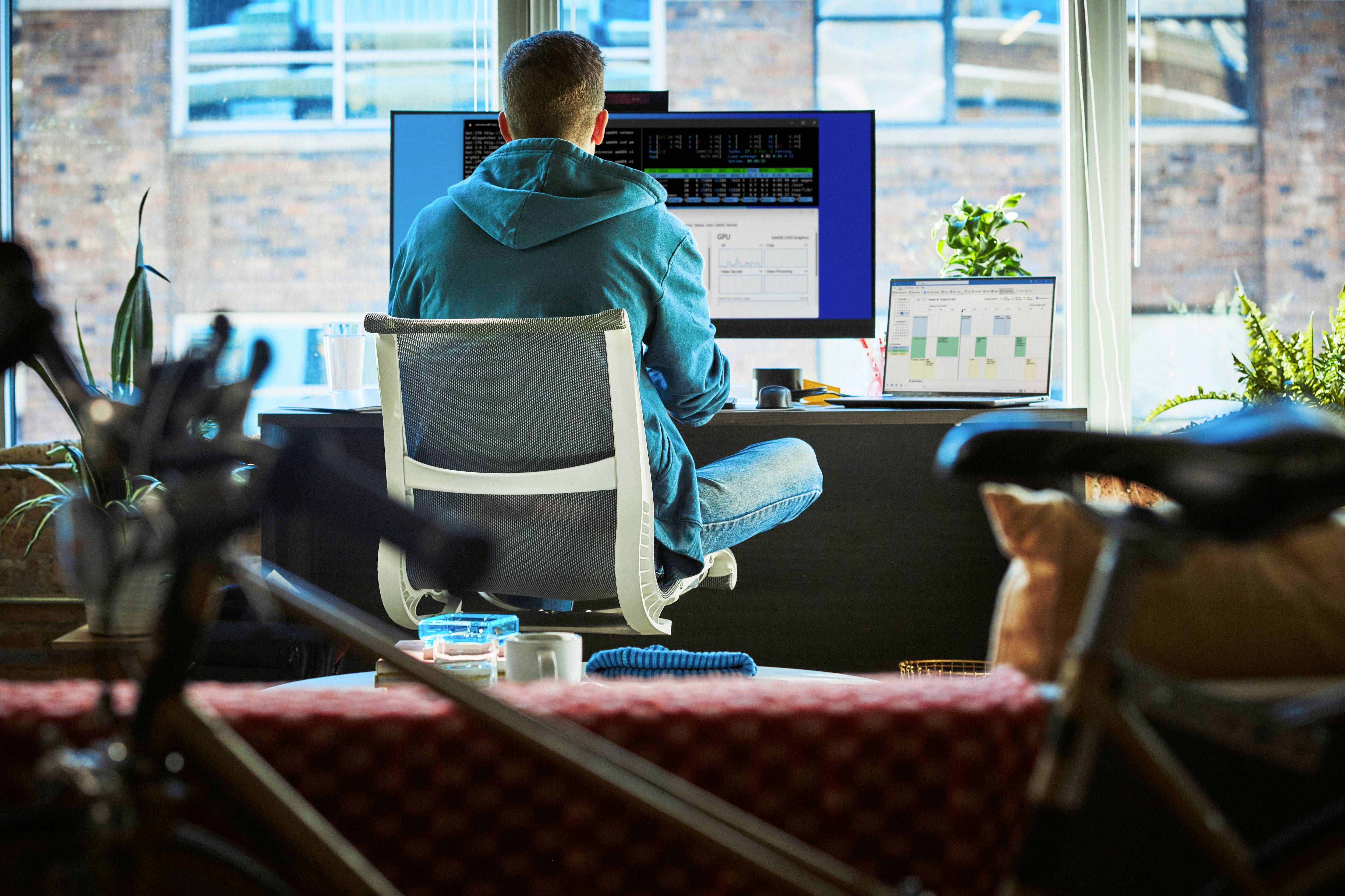 Ein Mann sitzt vor einem Computer und arbeitet in einem modernen Büro mit Glasfenster. Pflanzen umgeben den Schreibtisch.