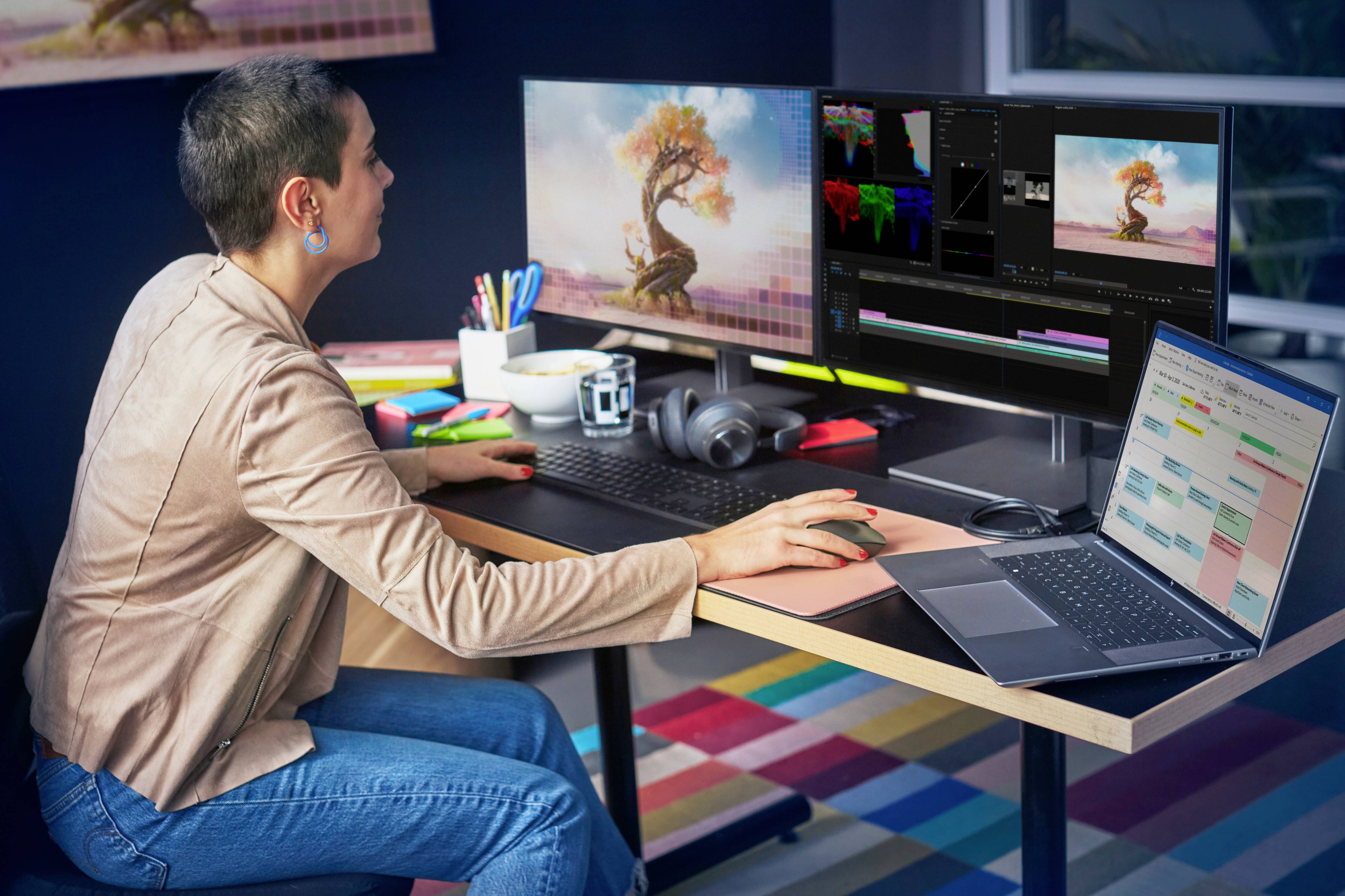 Eine Person bearbeitet ein Bild an zwei Monitoren, mit einem Laptop daneben. Der Raum ist modern mit einem bunten Teppich.