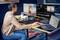 Eine Person bearbeitet ein Bild an zwei Monitoren, mit einem Laptop daneben. Der Raum ist modern mit einem bunten Teppich.