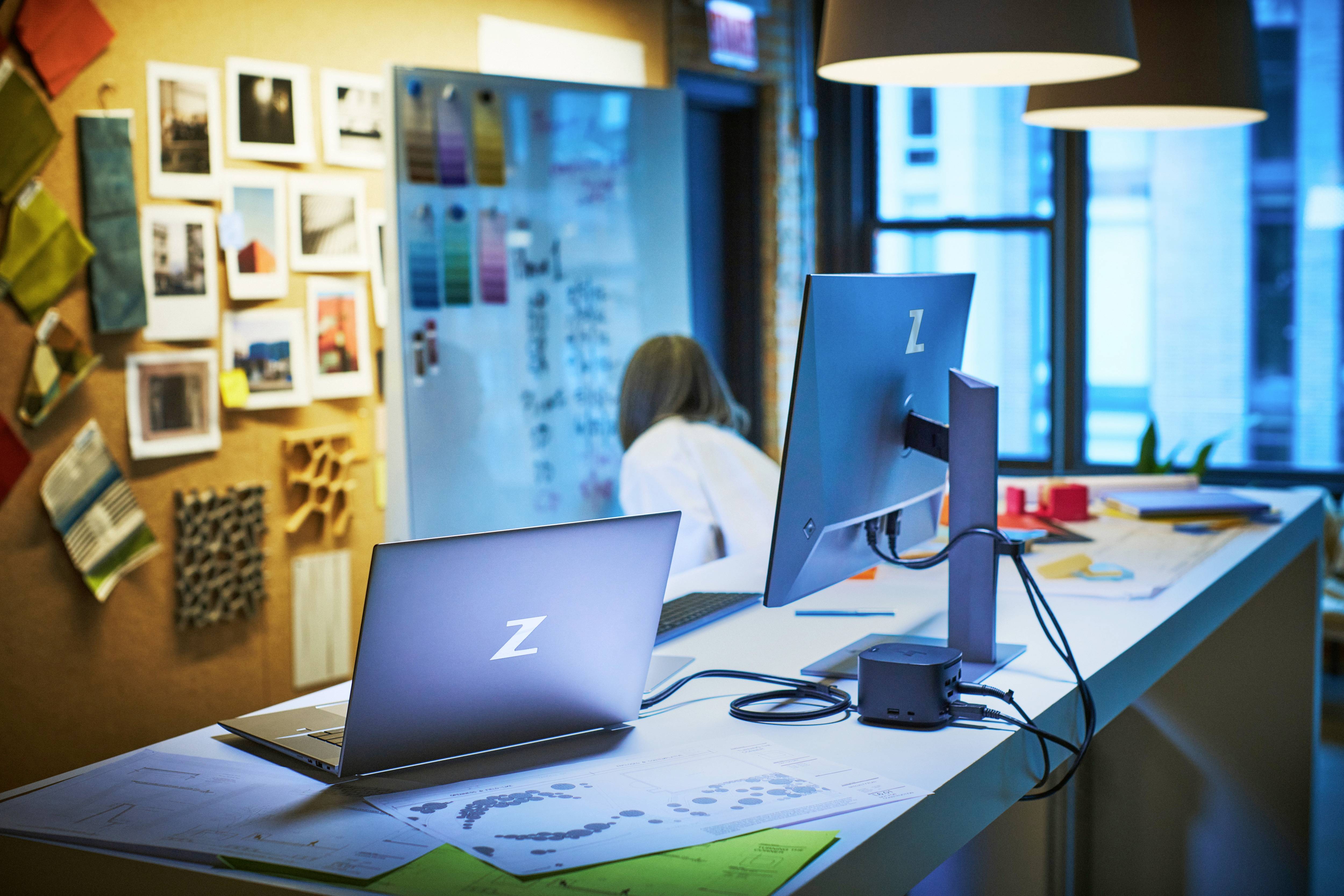 Ein modernes Büro mit zwei Laptops auf einem Tisch, im Hintergrund eine Pinnwand mit Mustern und eine Person, die an einem Whiteboard arbeitet.