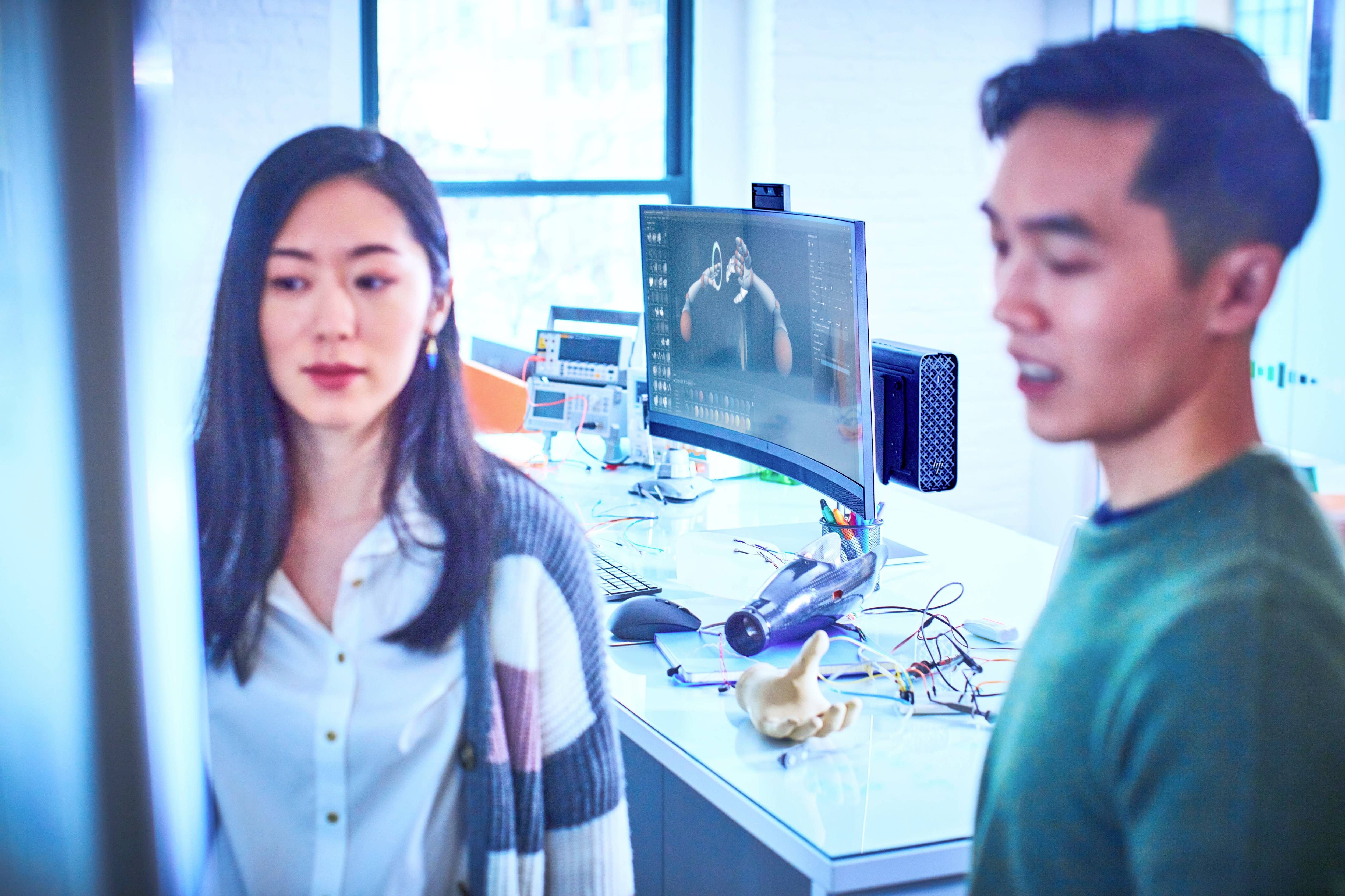Zwei Personen stehen vor einem Computerbildschirm in einem modernen Büro. Der Bildschirm zeigt eine technische Darstellung oder Animation. Im Hintergrund sind elektronische Geräte zu sehen.
