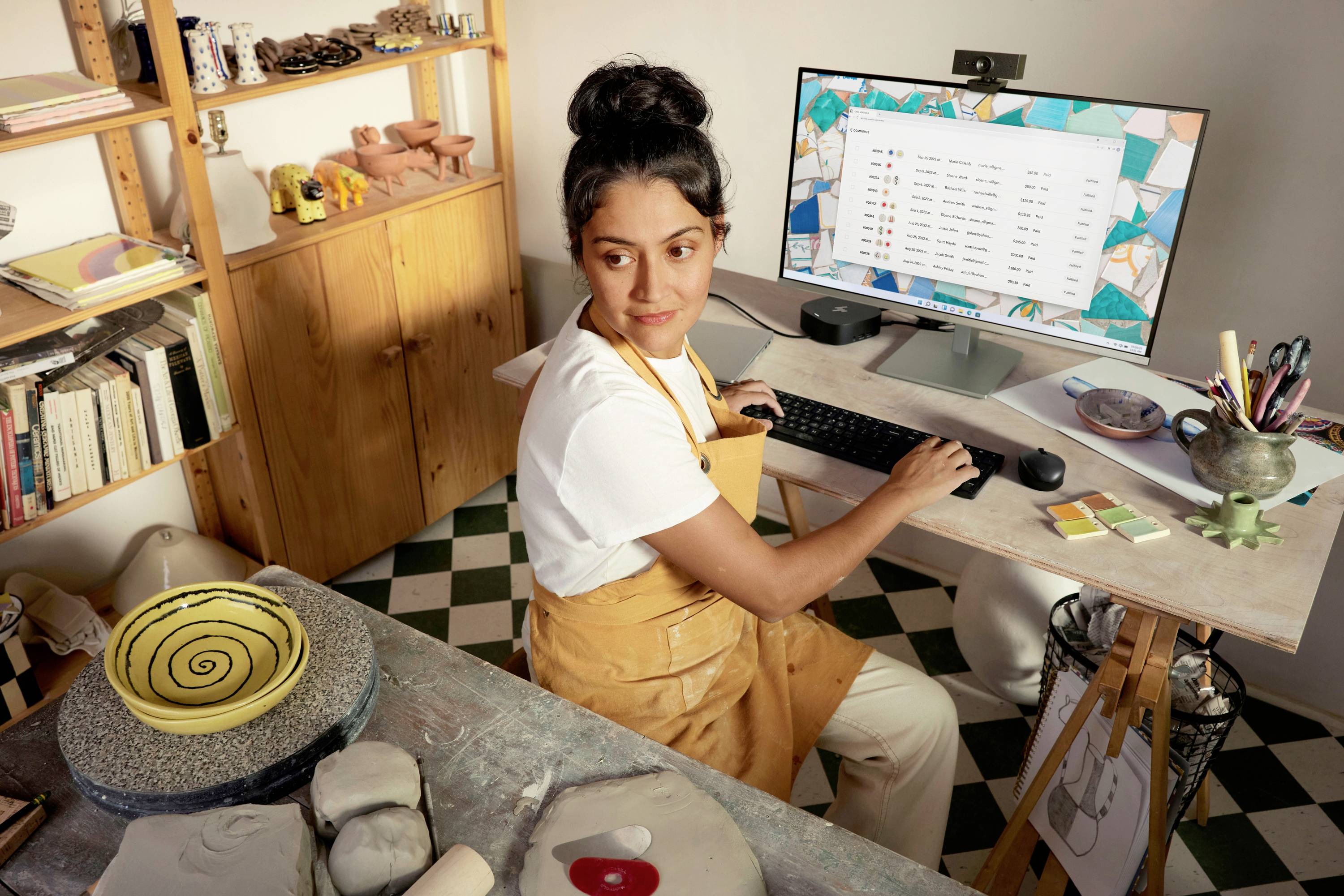 Eine Frau im Töpferstudio sitzt am Schreibtisch vor einem Computermonitor, der eine Dateiliste anzeigt, während sie sich nach rechts umschaut.