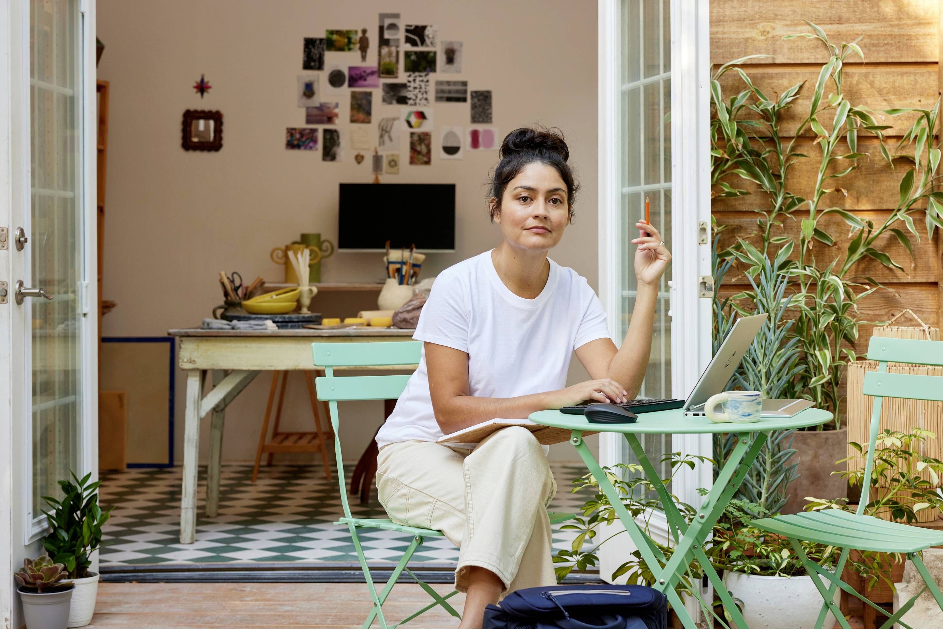 Eine Person sitzt an einem Laptop auf einer grünen Terrasse. Im Hintergrund ist ein gemütliches Zimmer mit einem Schreibtisch sichtbar.