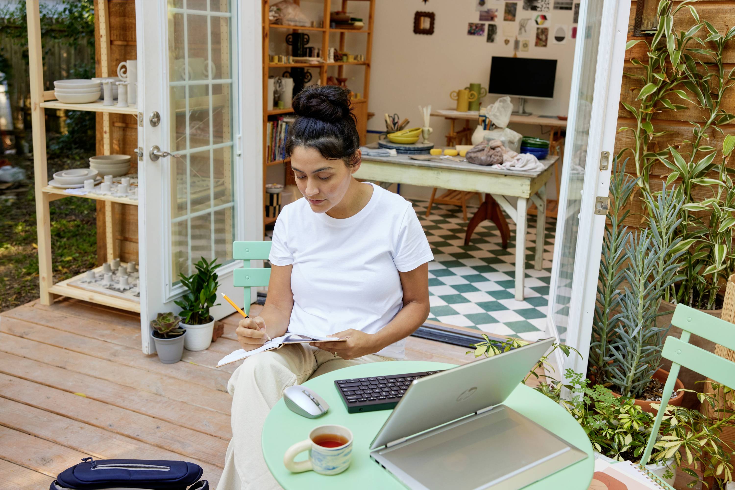 Eine Person sitzt auf einer Terrasse, schreibt in ein Notizbuch mit einem Laptop und einer Tasse Tee auf dem Tisch. Im Hintergrund ein helles Zimmer.